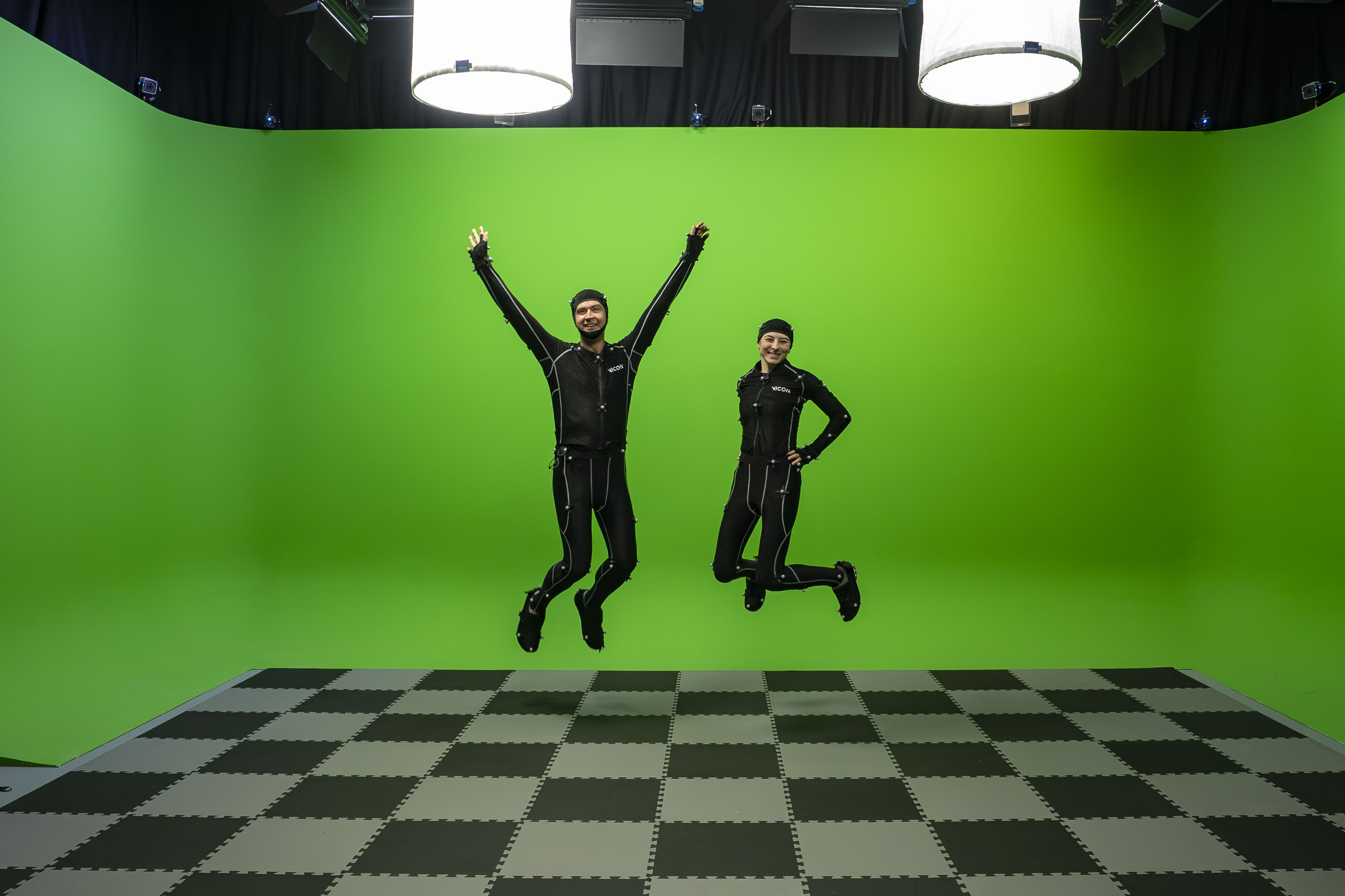 Two people wearing motion capture suits jumping in front of a Chroma-Key green surround.