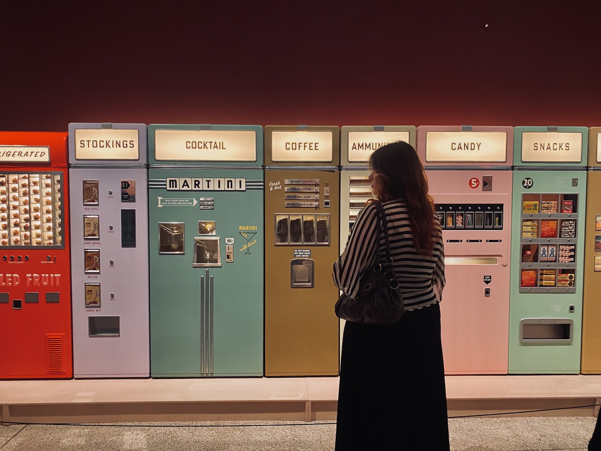 Person in striped top stands before row of colourful vintage vending machines including red, white, green, and pink units against dark red wall.