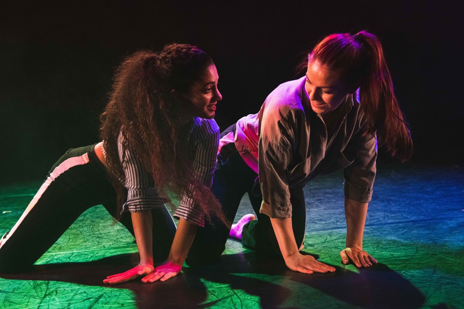 Two women kneel on a stage in a mirrored floor exercise pose under pink and green lighting.