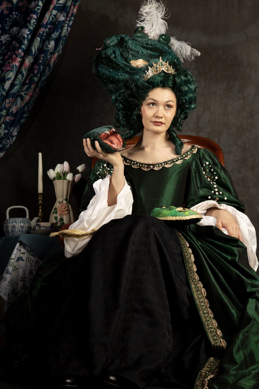 A woman with elaborate green hair styled high, wearing a dark green gown adorned with beads and lace, sits in a chair. She holds a dessert on a plate in her right hand, with a table set behind her displaying candles and porcelain.