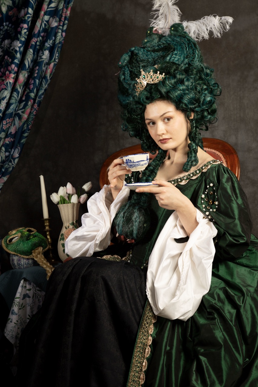 A woman dressed in a green historical gown with puffy sleeves sits on a chair, holding a teacup. Her elaborate hairstyle is adorned with a tiara and feathers. A patterned curtain and a small table with a teapot and flowers are visible in the background.