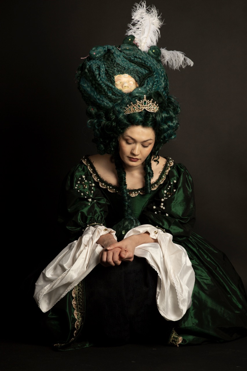 A woman wearing a green historical costume with an elaborate hairstyle sits with her head bowed. The outfit includes intricate details and a white underskirt, while the background is dark, enhancing the focus on her attire.