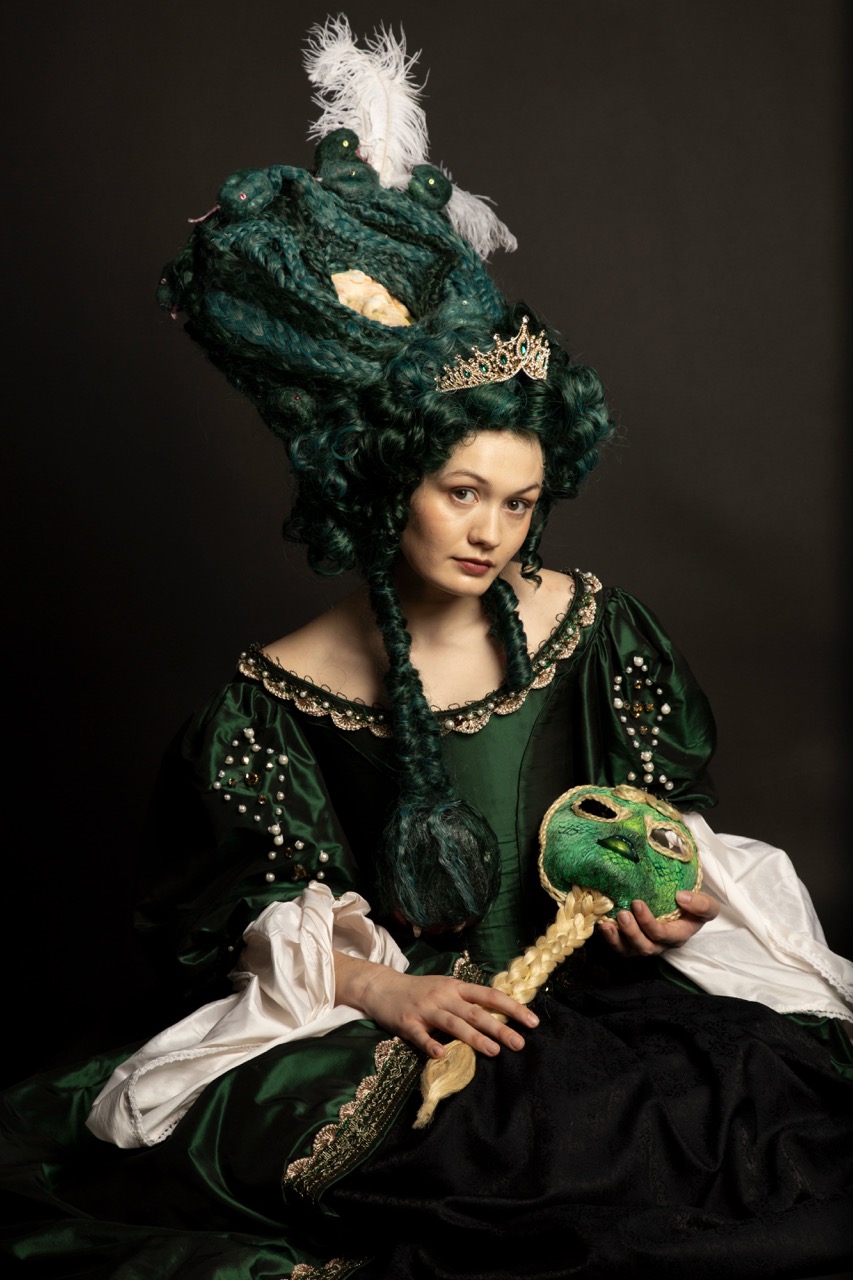 A woman dressed in a green historical gown with decorative embellishments sits against a dark background. Her elaborate hair, styled in braids and adorned with feathers, complements her regal appearance. She holds a green frog-like object in one hand.