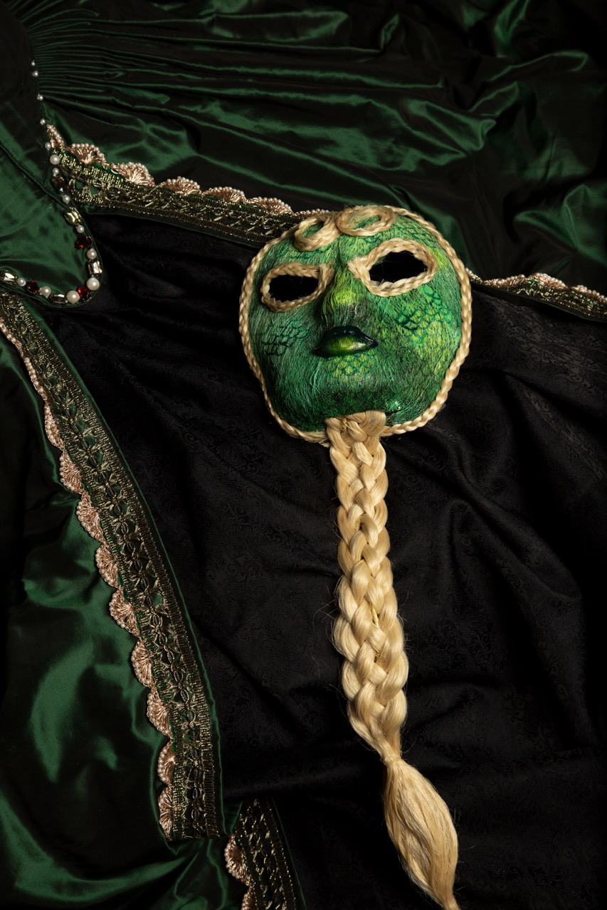 A green mask with intricate designs and a braided blonde hair extension is placed on a dark fabric adorned with green silk. The surrounding fabric features a decorative border with a metallic sheen, enhancing the mask's ornate appearance.