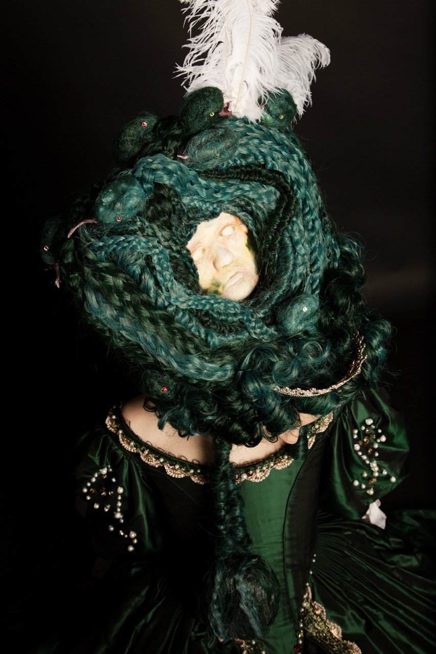 A person wearing an elaborate dark green gown with intricate gold detailing. Their hair is styled into large twisted braids, and a white feathered ornament is positioned atop the head, creating a dramatic appearance against a black background.