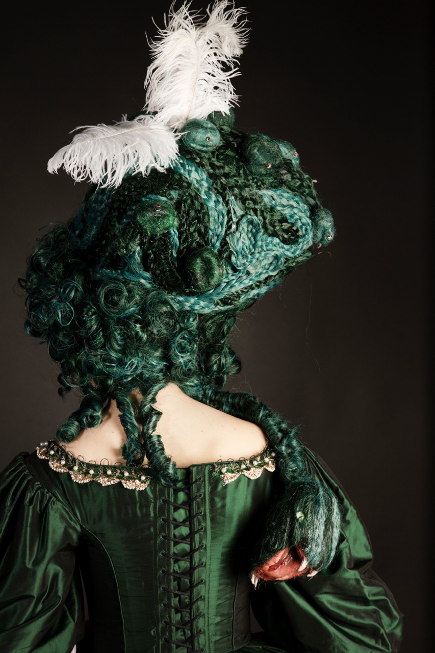 A person wearing a dramatic green dress with a fitted bodice and puffed sleeves, showcasing intricate rope-like hairstyles adorned with white feathers. The figure is shown from behind against a dark background.