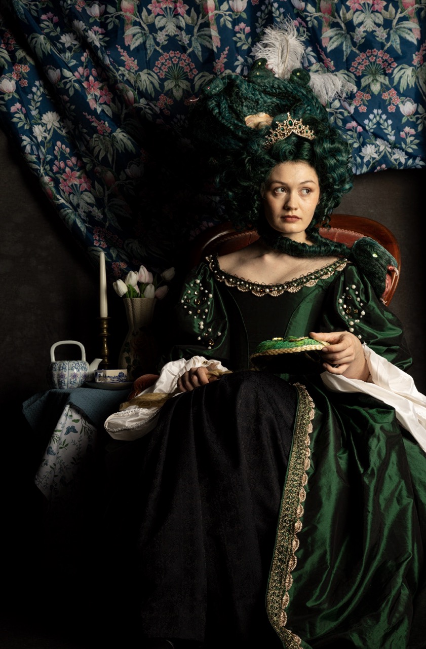 A woman in an elaborate green gown sits in an armchair, holding a dessert. The gown has intricate detailing and puffed sleeves, while a large, ornate hairpiece adorns her dark hair. Behind her is a patterned fabric backdrop.