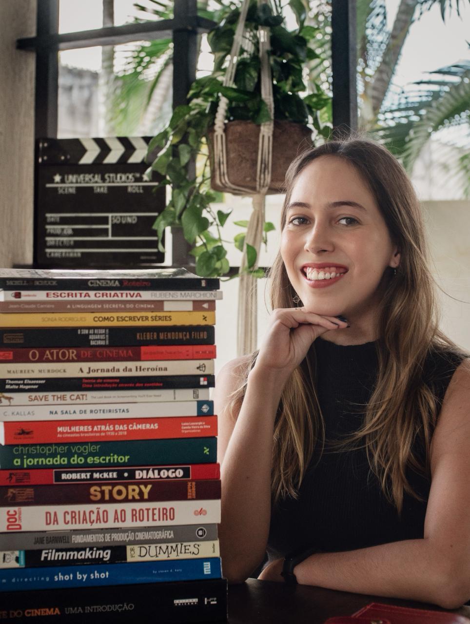 Smiling woman with long brown hair sits beside tall stack of filmmaking books, with film clapperboard and hanging plant visible.