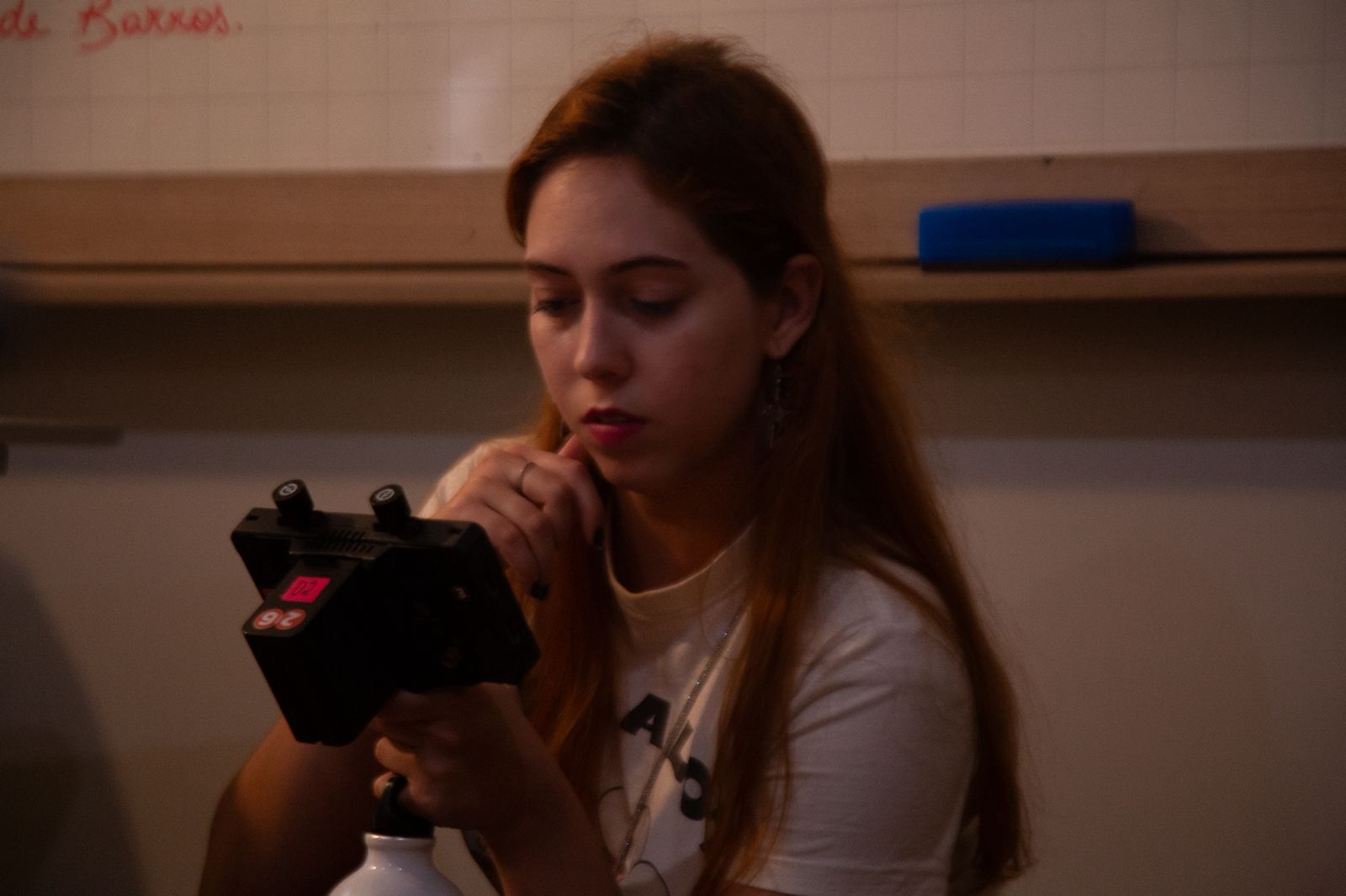 Woman with long brown hair looking down at black camera in kitchen setting with white cabinets and shelving visible behind her.