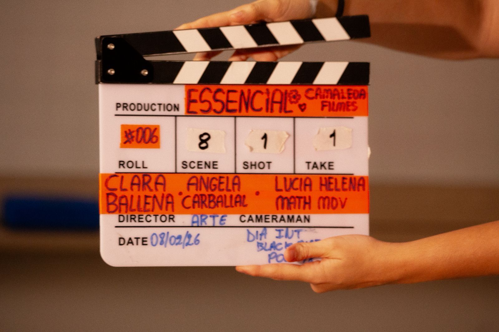 Film clapperboard held by hands showing "Essentials" production details with black and white striped top, white body with red text.
