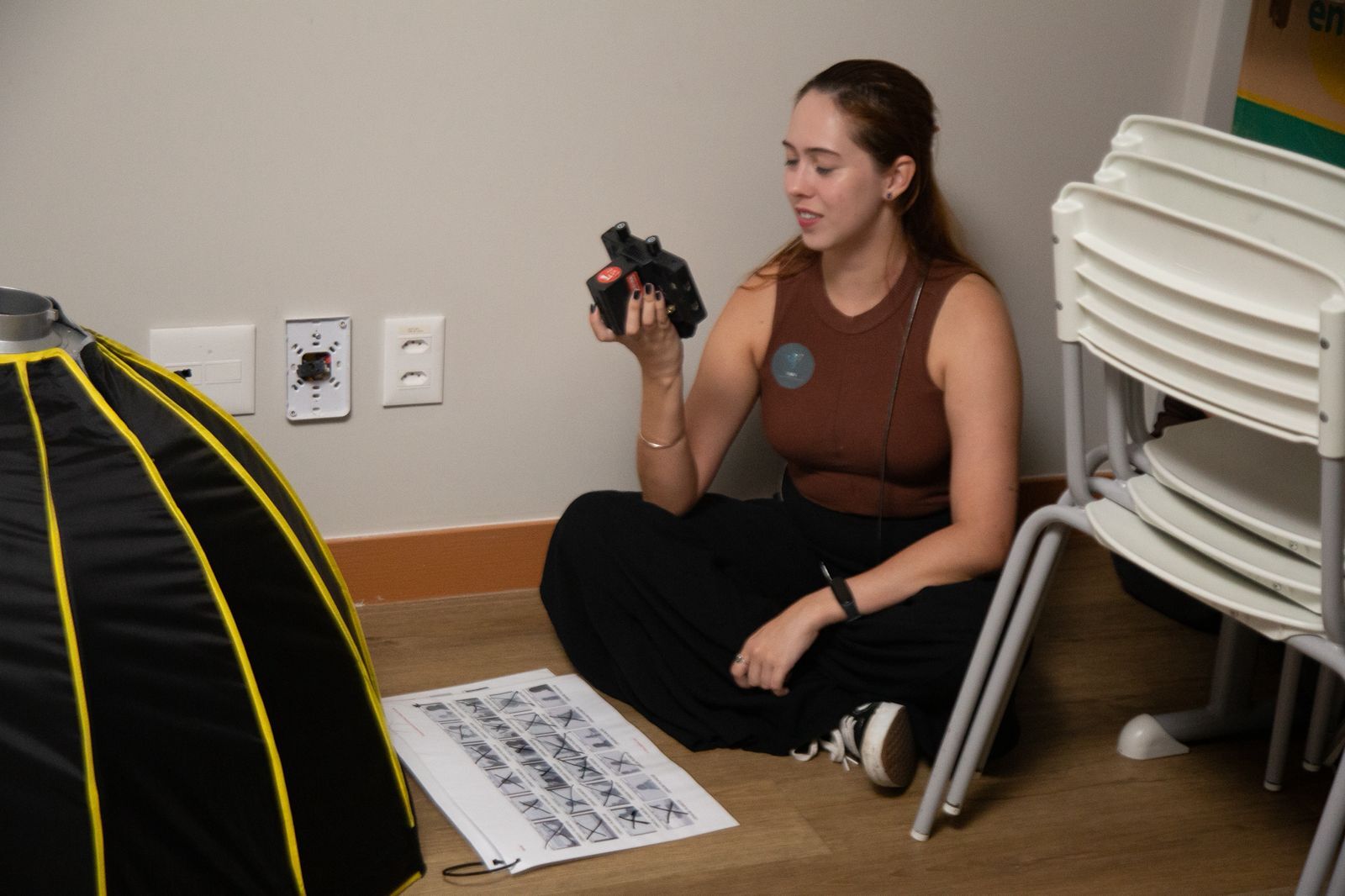 Woman in brown vest sits on floor holding camera, with stacked white chairs and yellow-striped black object nearby.