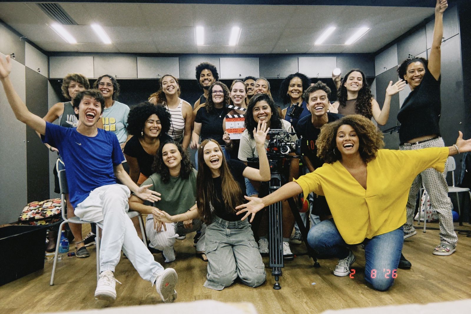 Group of young people celebrating in dance studio, arms raised, smiling and cheering. Woman in bright yellow top prominent in foreground.