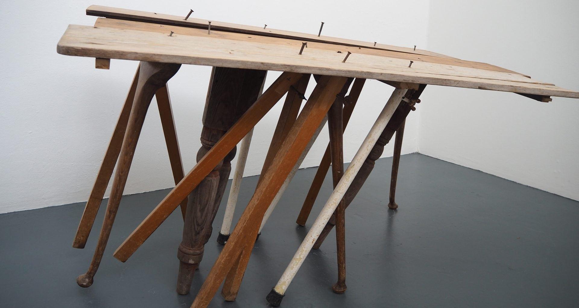 Sculpture made from assorted dining room table legs and three planks of wood roughly nailed together.