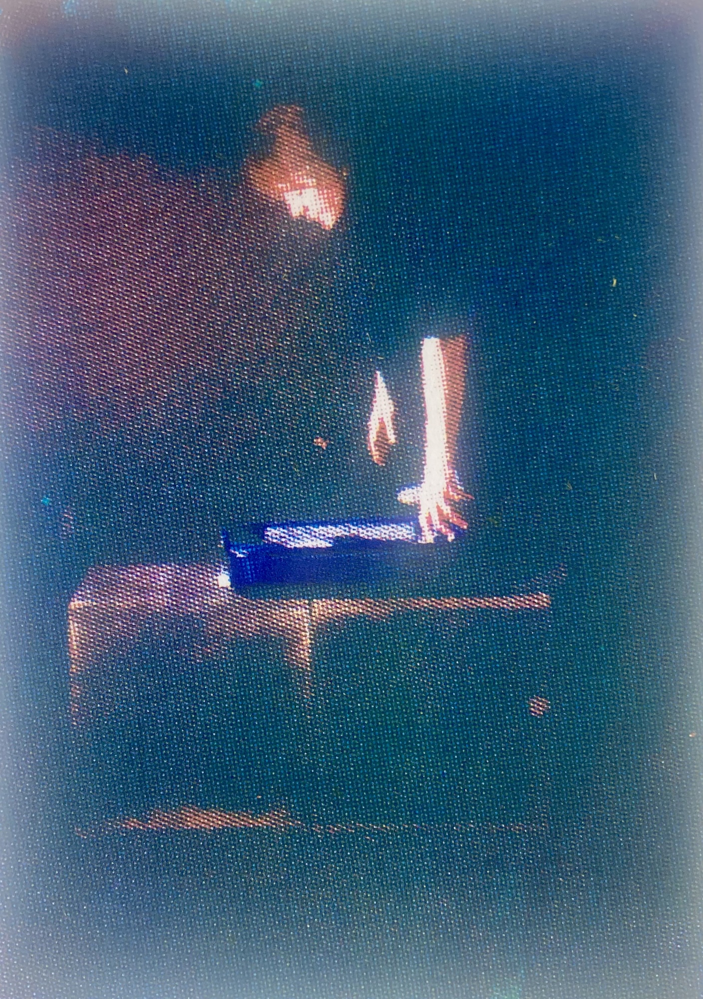 A dimly lit scene featuring a person with long hair positioned over a blue object on a table. The background is dark, enhancing the focus on the figure and the object being handled.