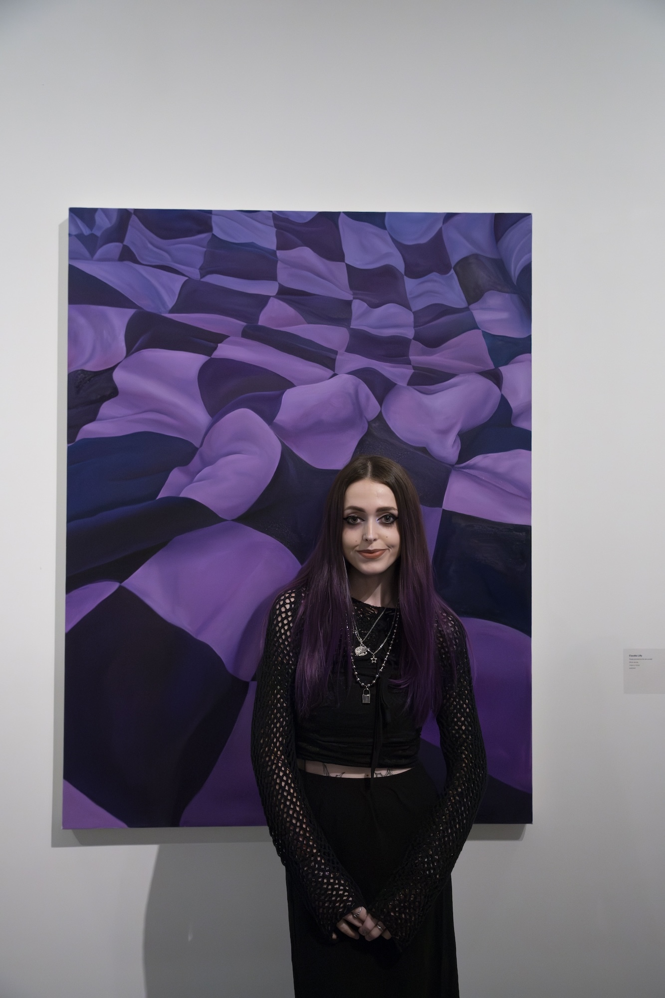 Artist C Lilly standing in front of a canvas of their painting, showing purple and indigo checked fabric.