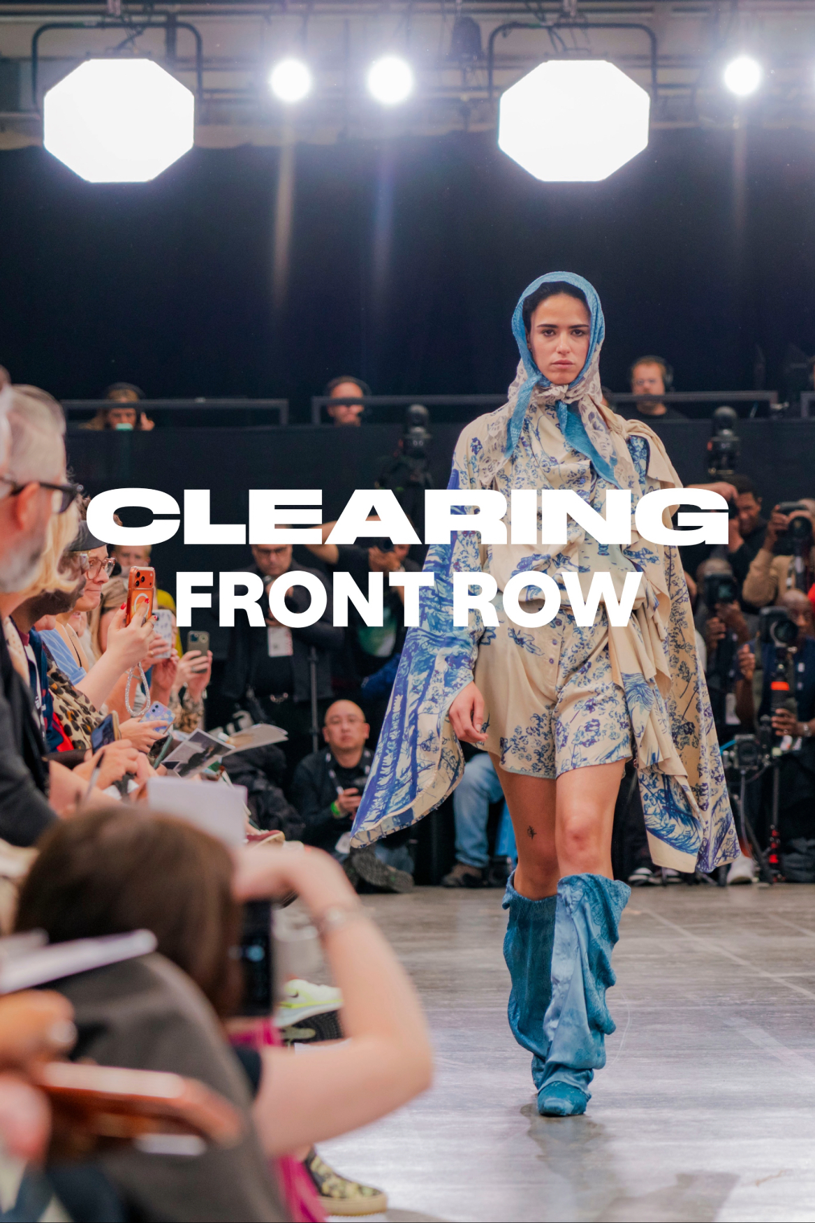 A model walks down a runway wearing a patterned outfit with a hood, while attendees in the front row take photos. The scene is set in a fashion show with bright lights and a crowd observing the presentation.