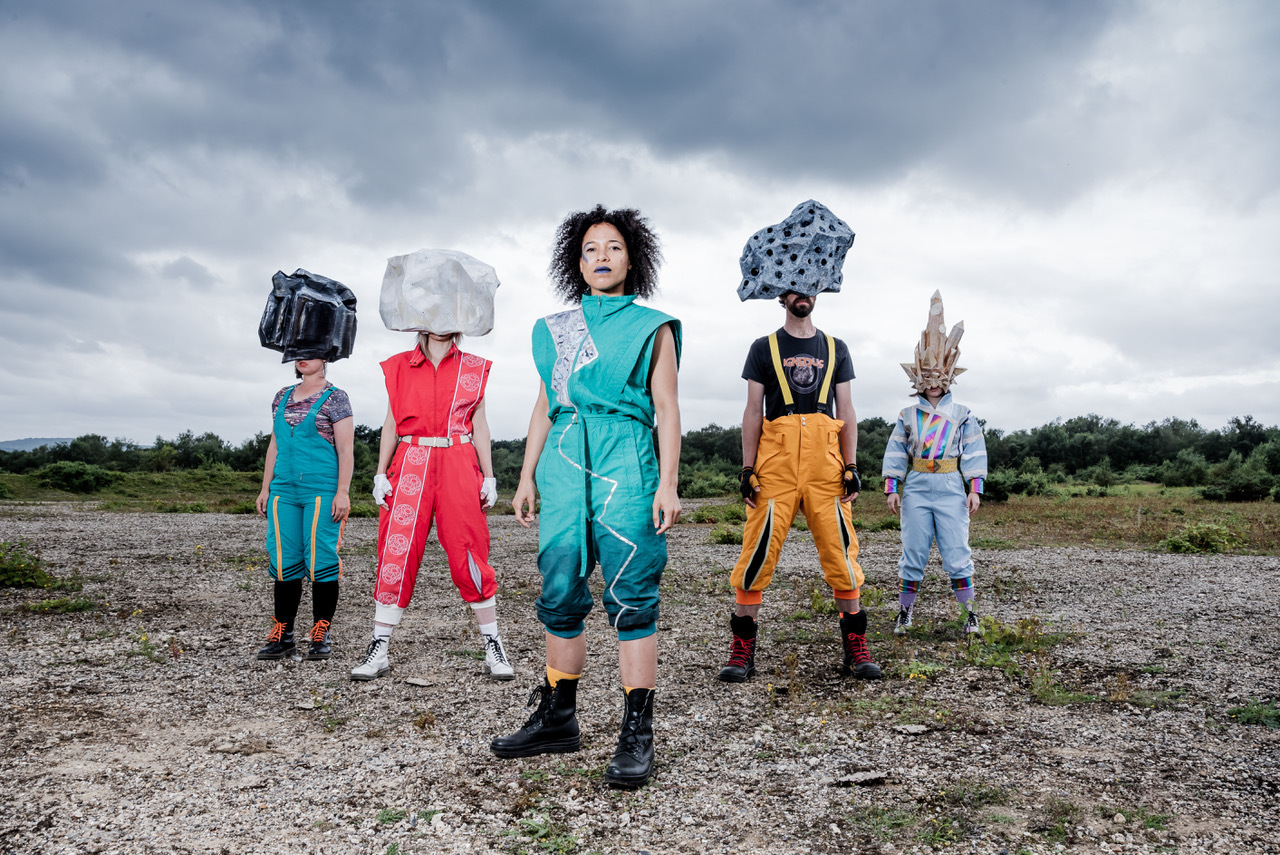 Five people stand in a triangle formation wearing different costumes. Four of them are wearing headdresses shaped like different rocks.