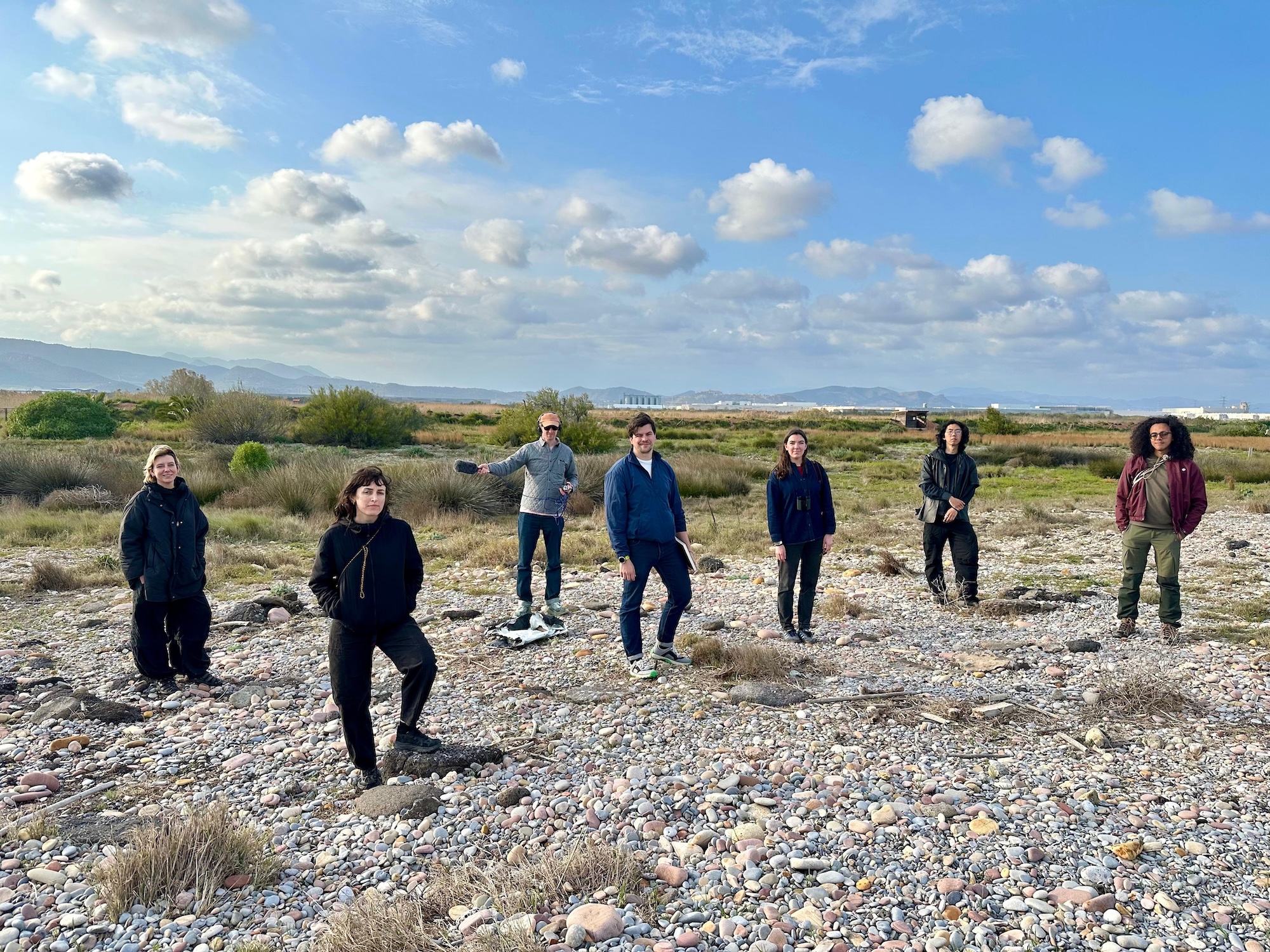 A group of seven people standing on a rocky area beside a riverbank. They are arranged in a semi-circle, with greenery and a blue sky dotted with clouds in the background.
