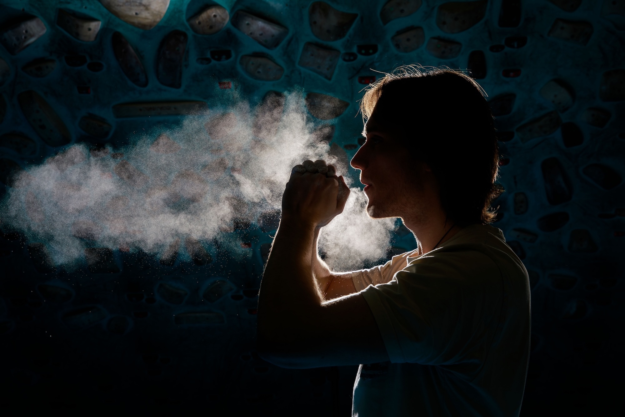 Climber viewed from the side in low lighting with hands raised. Chalk dust floats in the air in front of them. A climbing wall is dimly visible in the background.