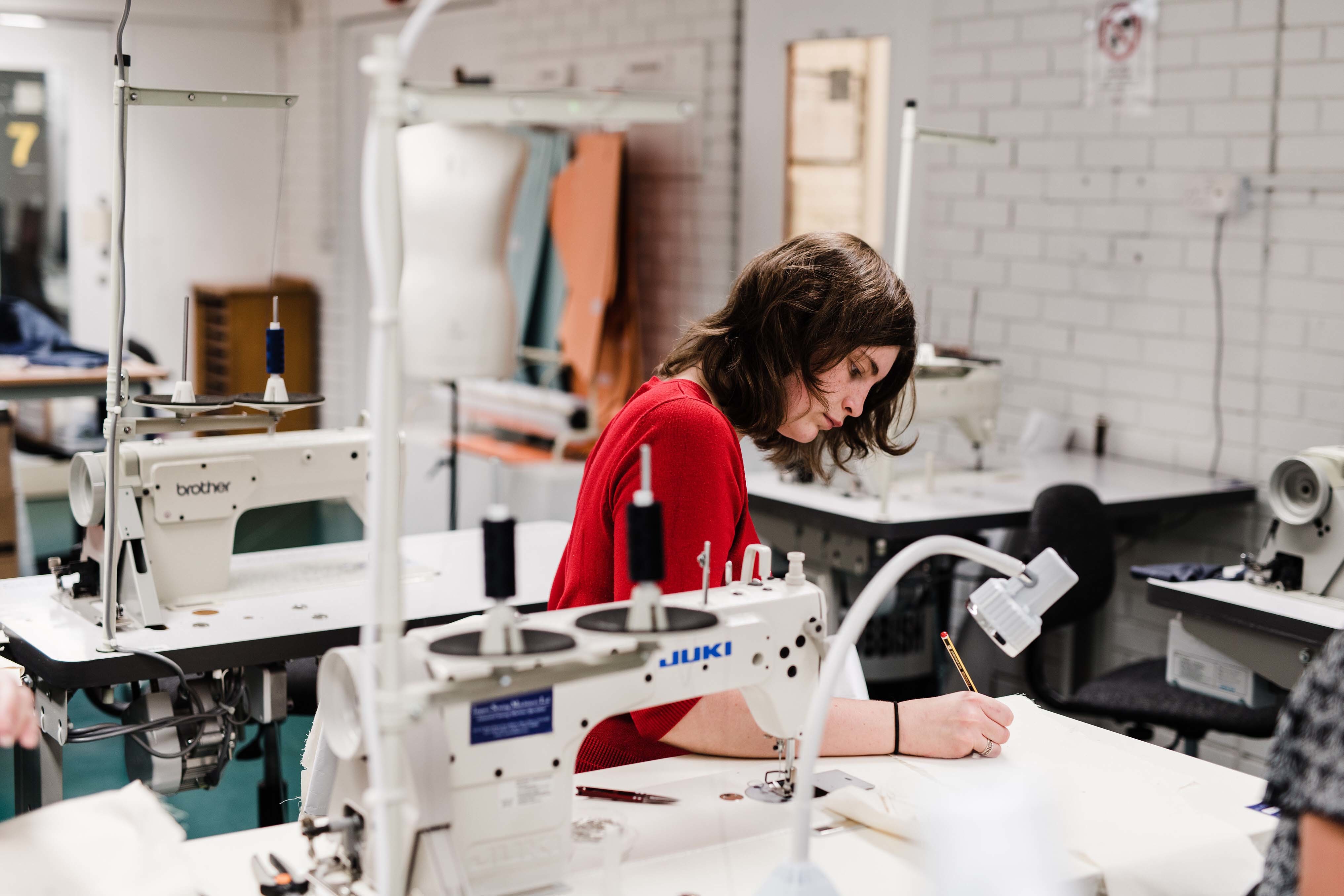 Person sits at a table with a sewing machine on it and draws on a piece of paper.