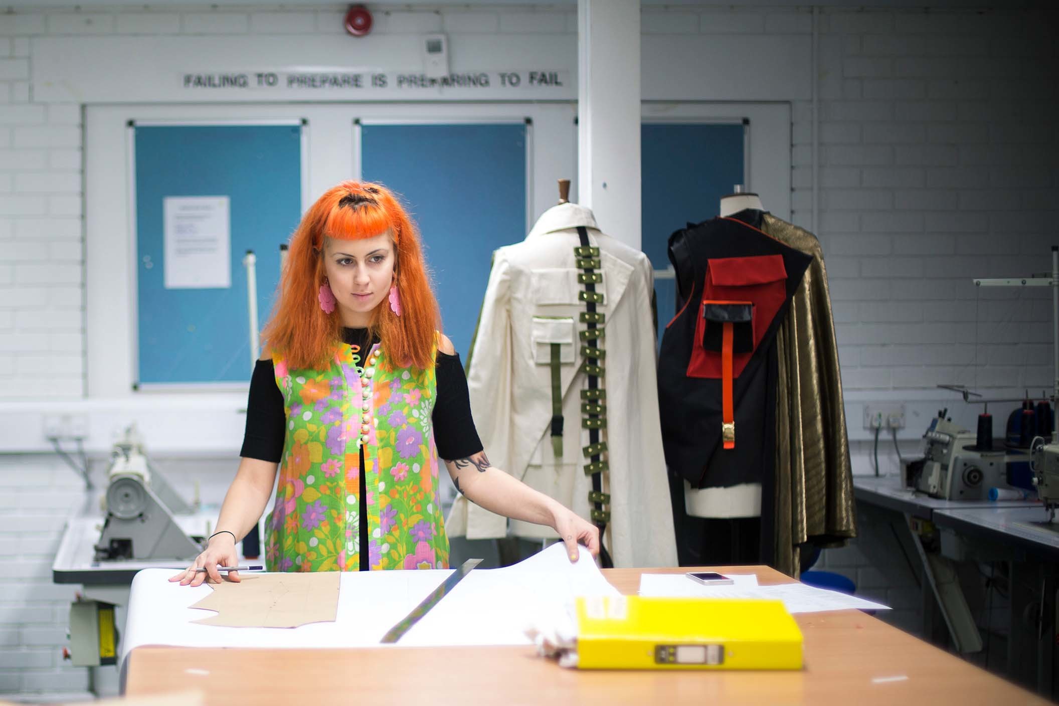 A person stands at a table with a large piece of paper unrolled in front of them. There are two dressed mannequins in the background.