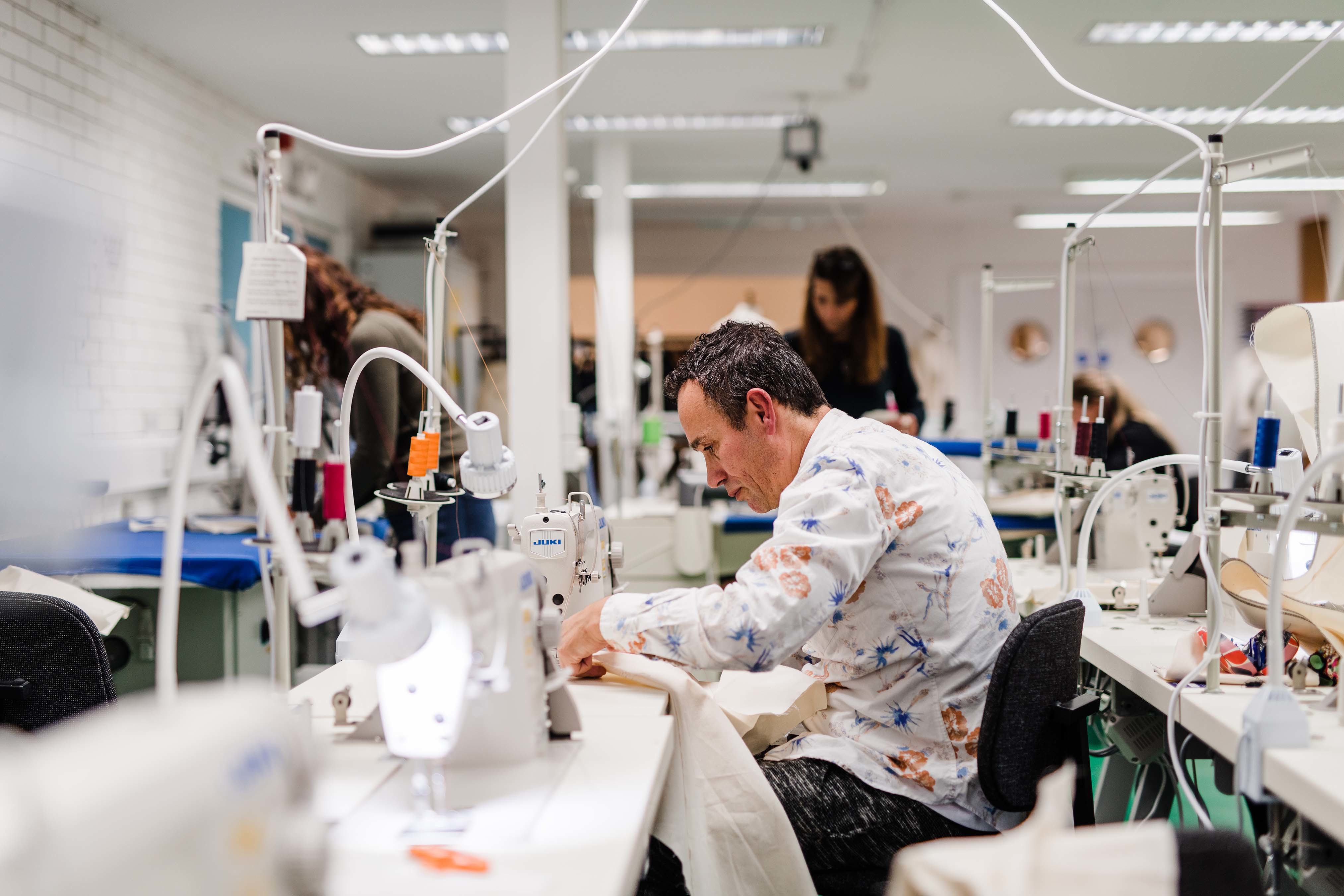 A person sits in a costume studio at a bank of sewing machines and works on a piece of fabric.