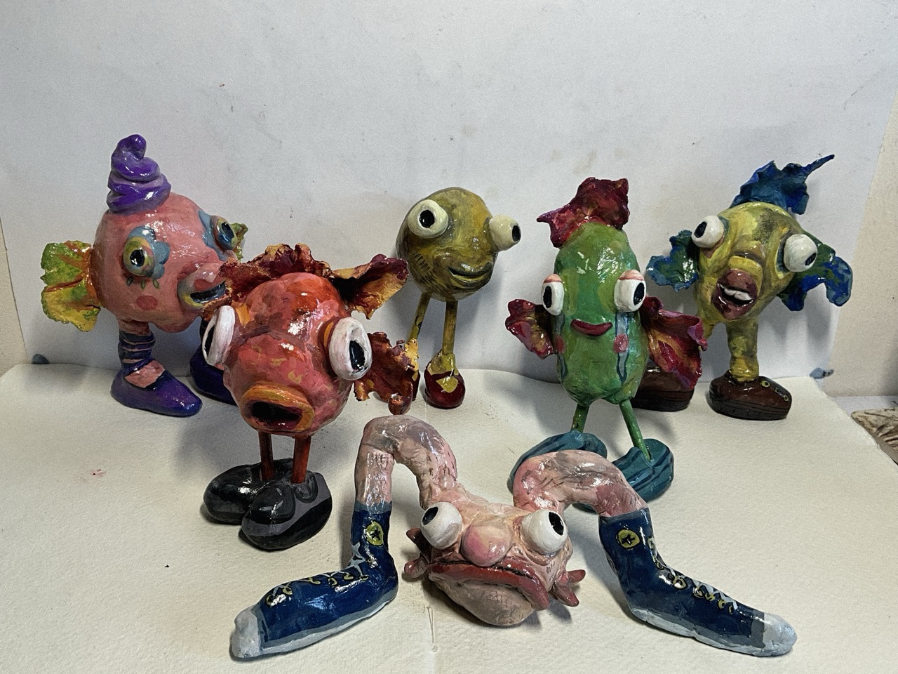 Several colourful handmade figurines with oversized eyes are arranged on a white surface against a plain wall, including fish-like characters and a pink creature with blue shoes in the foreground.