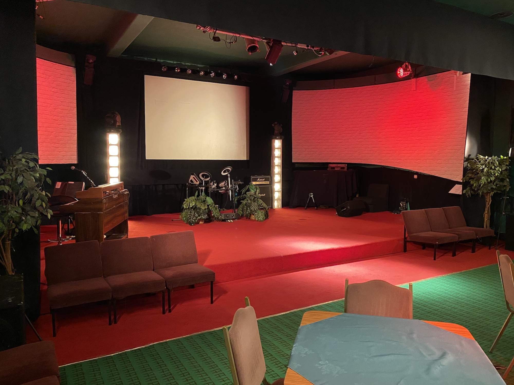 Empty stage in the Club Room at the Palace Court Theatre, with a piano on the left side, drums and a screen in the background, two blocks of chairs facing the camera in the foreground.