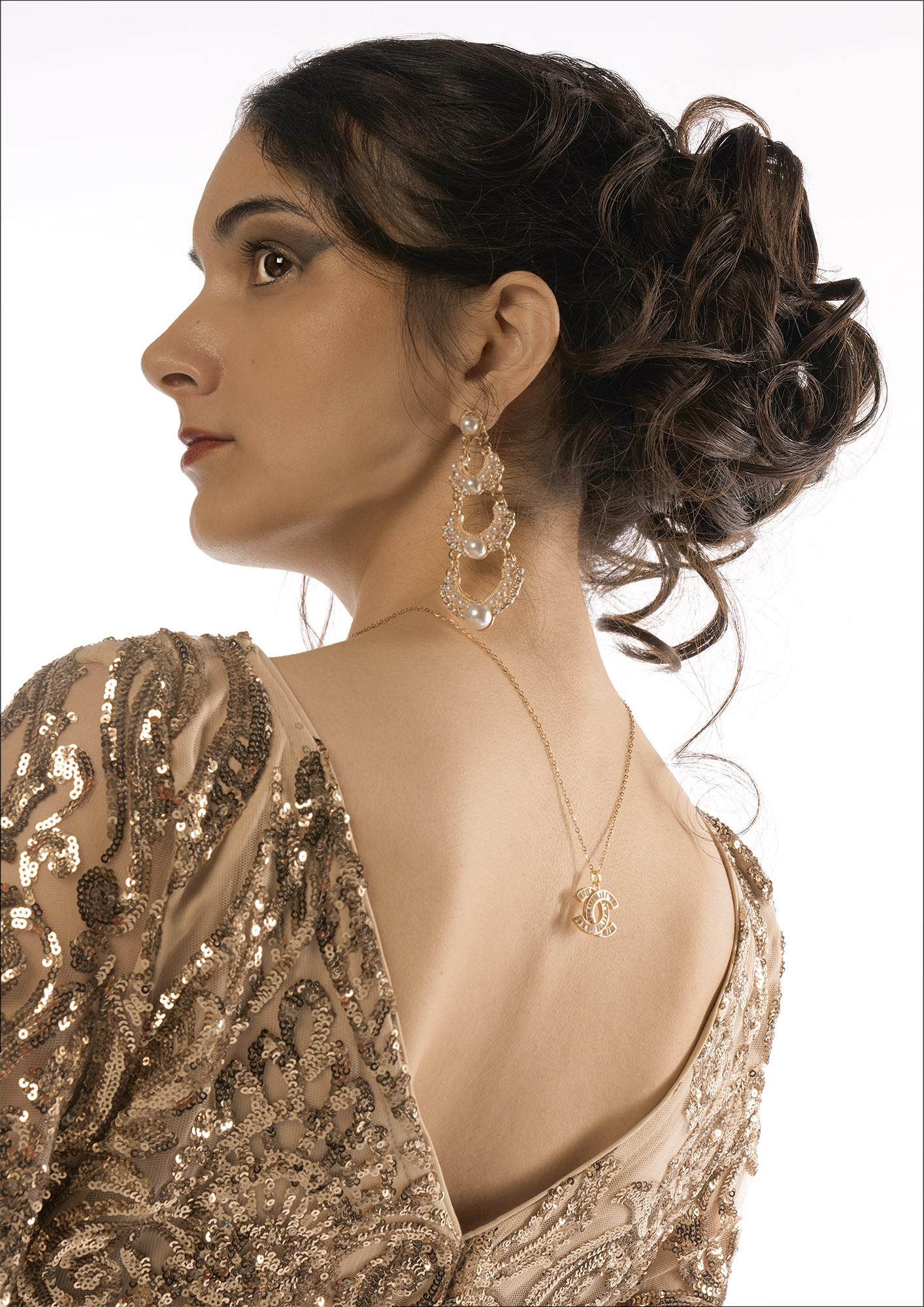 A woman in a gold sequinned dress is shown in side profile with an updo hairstyle and long earrings against a plain white background.