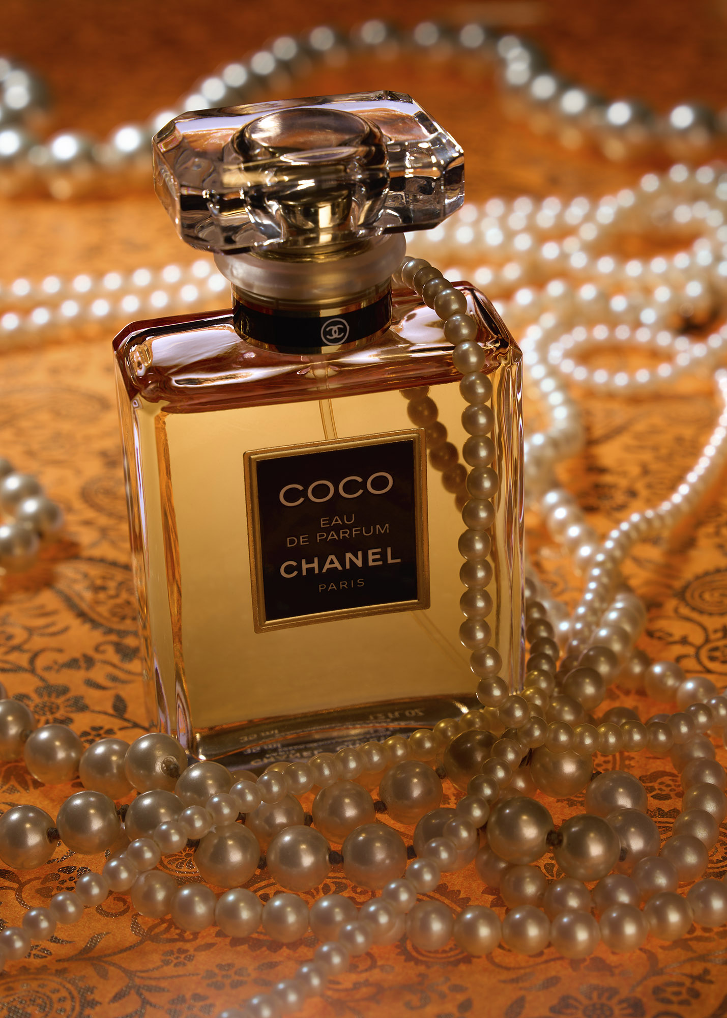 A Chanel Coco Eau de Parfum bottle with pearl strands draped around it on an orange patterned surface.