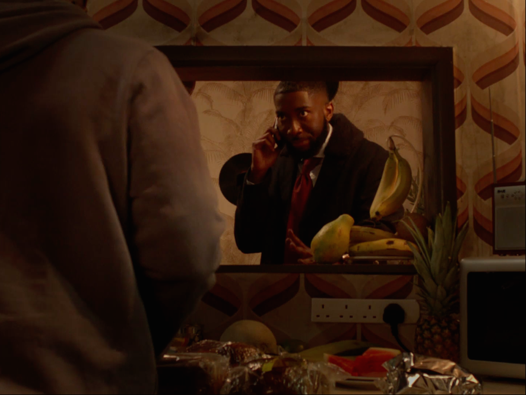 Film still of a man on the phone in a suit, reflected in a mirror, 70s-style wallpaper.