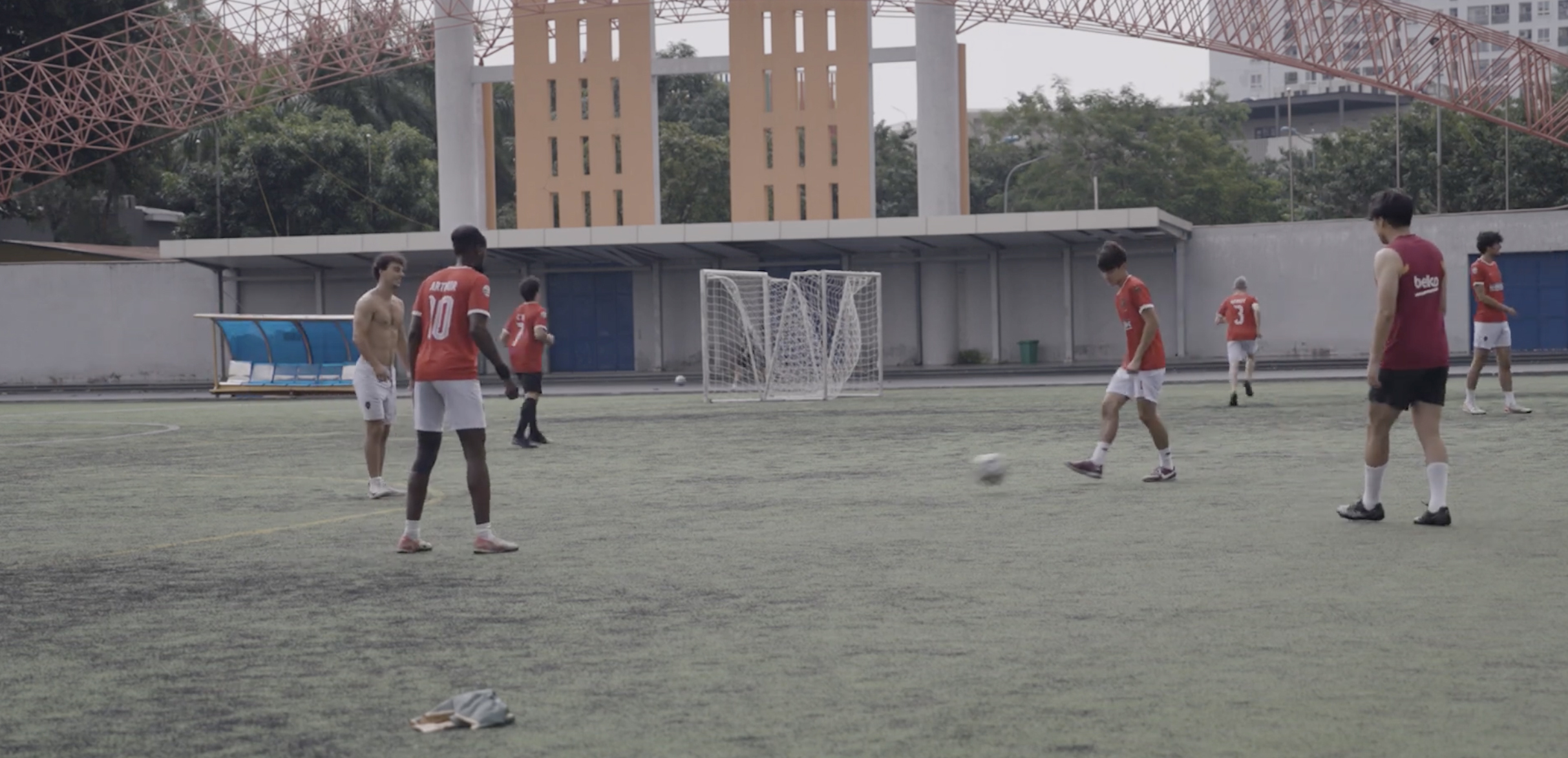 Football players in red and white shirts on outdoor pitch with goal posts, buildings and bridge visible in background.