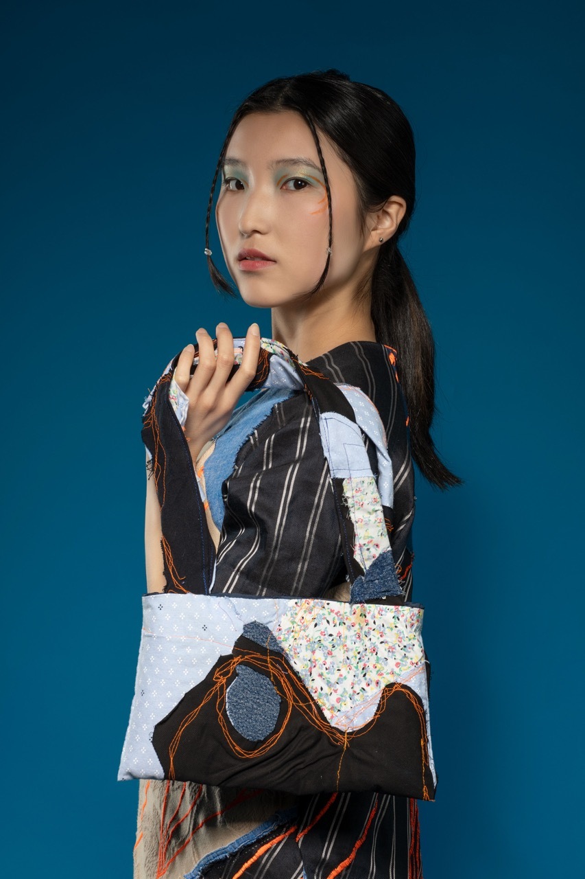 Woman in patchwork dress with black, blue and white fabric, holding a handbag up, deep blue background.