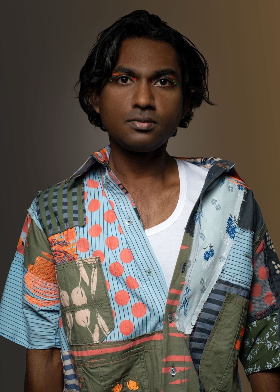 Person with dark hair wearing colourful patchwork overshirt with mixed patterns including stripes, polka dots, and floral prints over white top.