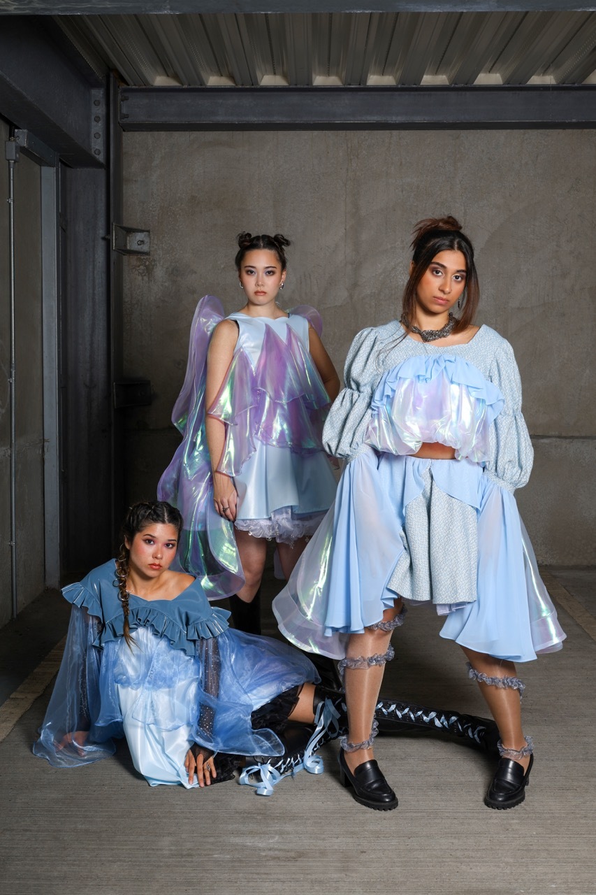 Three women in iridescent blue garments in industrial concrete space with exposed ceiling beams and metal framework.