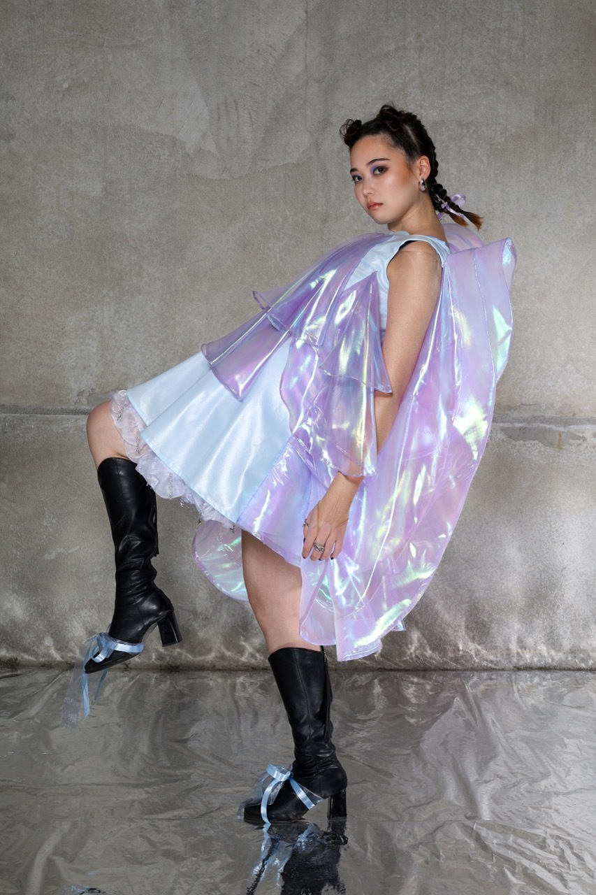 Woman in iridescent purple cape-style garment and black knee-high boots posing against grey concrete wall and floor.