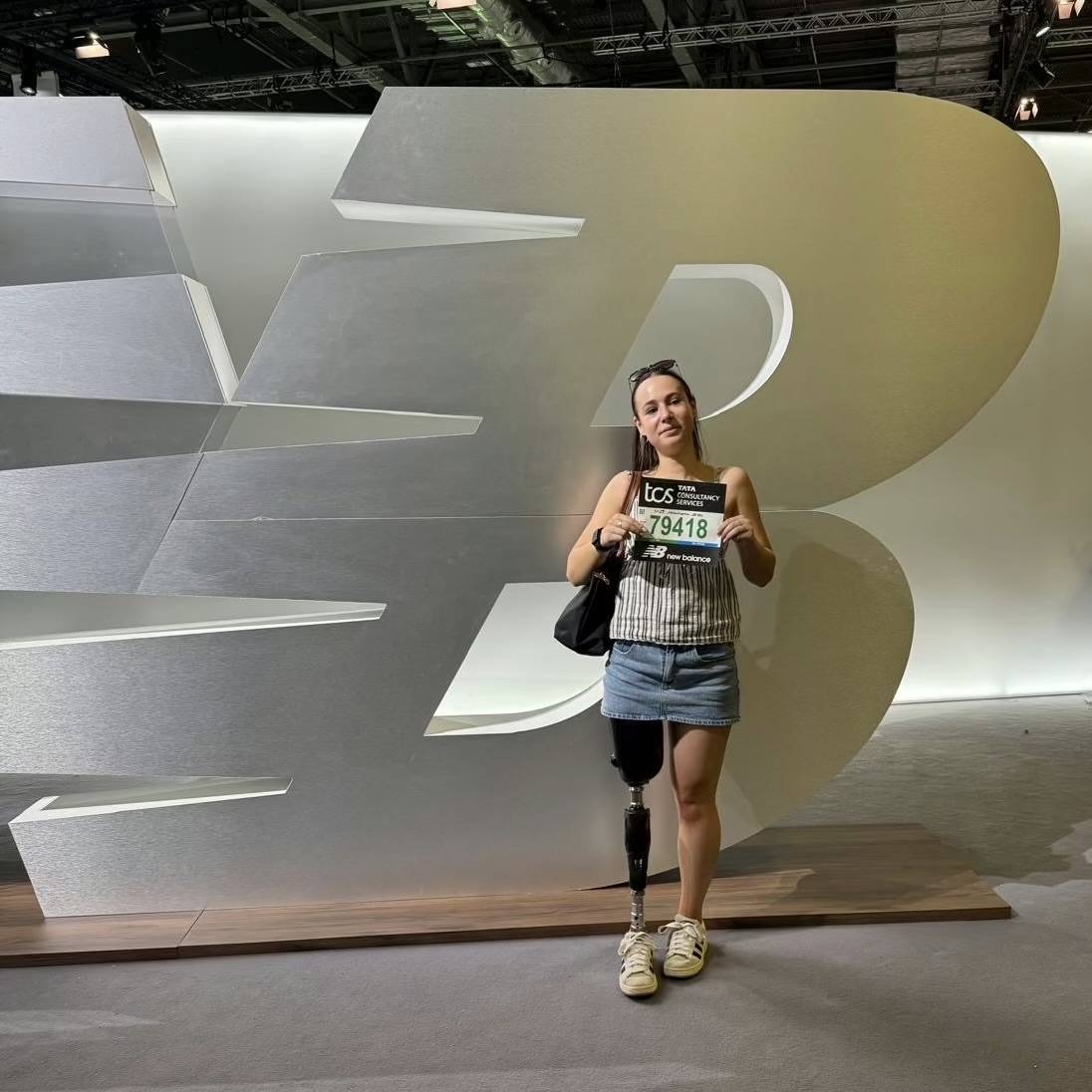 Amelia Mason holding her marathon number stood in front of a large, white New Balance logo, illuminated in the background.