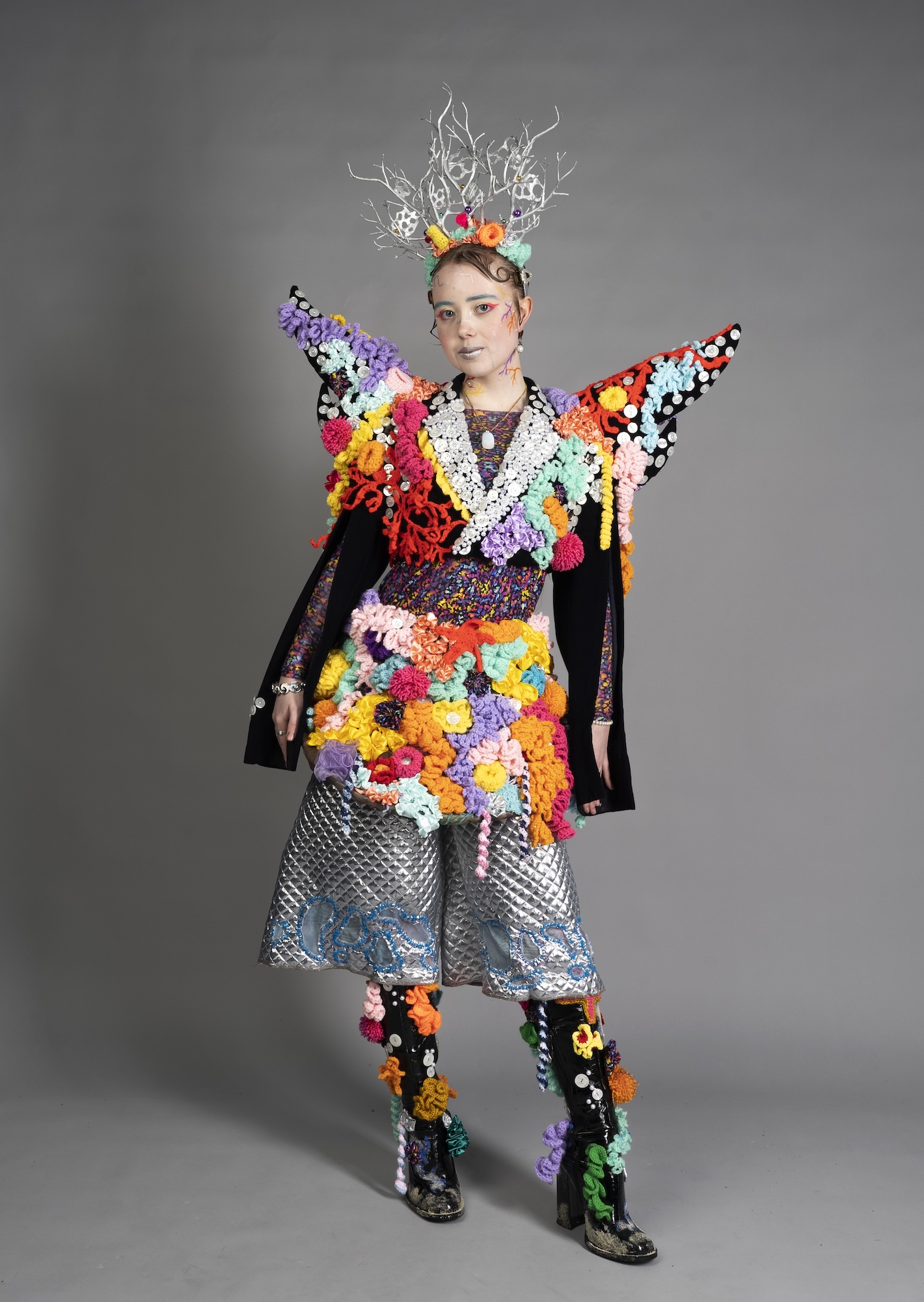 A model wearing a colourful avant-garde outfit with floral appliqués, a silver patterned skirt and black boots, posed against a plain grey studio background.