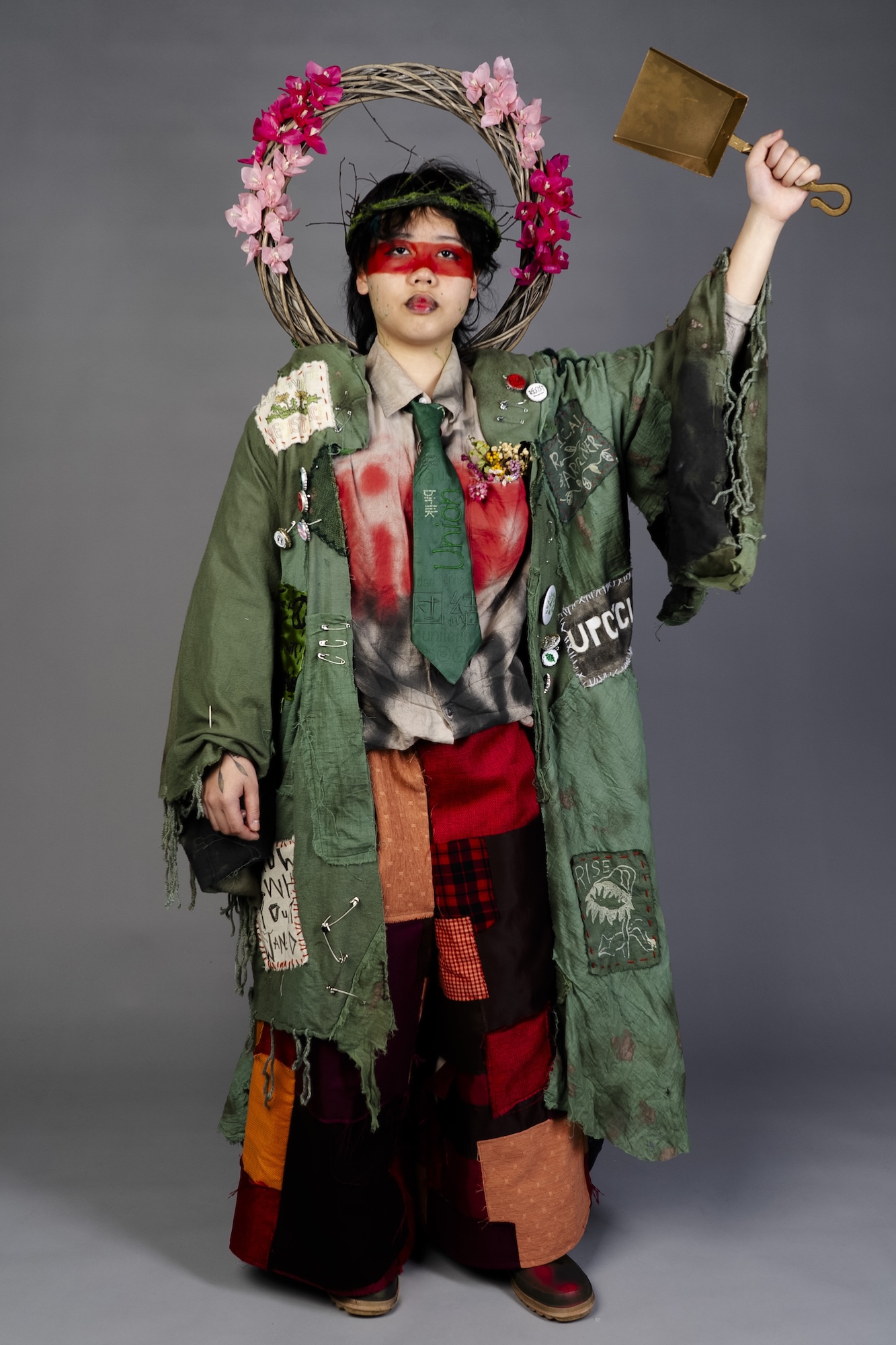 A person in a patchworked green outfit with a floral wreath and red eye covering holds up a brass bell against a grey studio background.