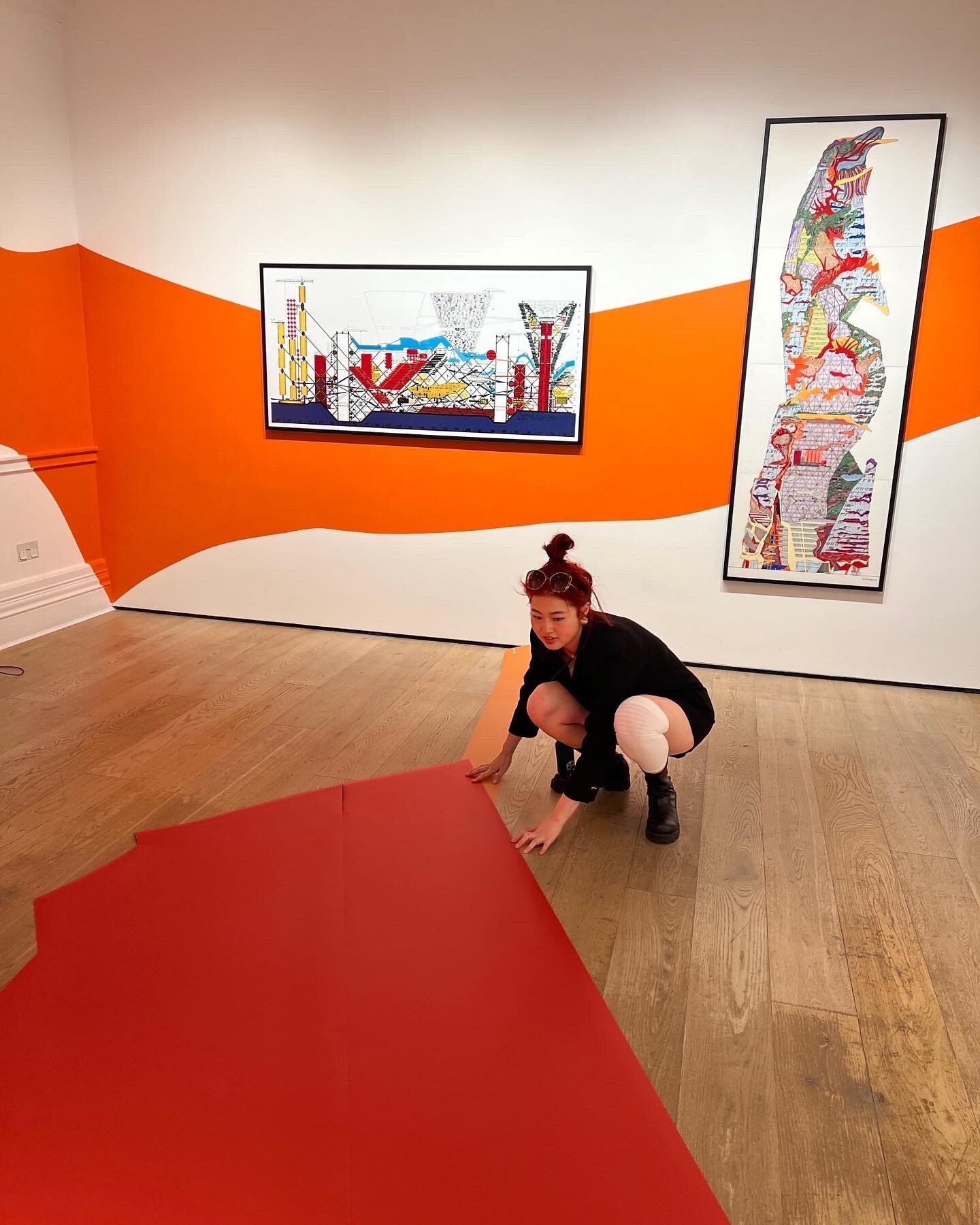 C Ding in gallery space with orange geometric wall design, red floor section, and two framed artworks.