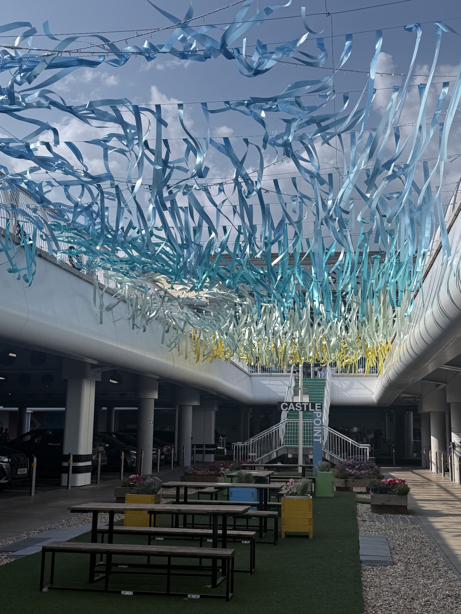 A shopping centre courtyard with blue ribbon streamers overhead, picnic benches and a green sign reading Castle Plaza in the centre.
