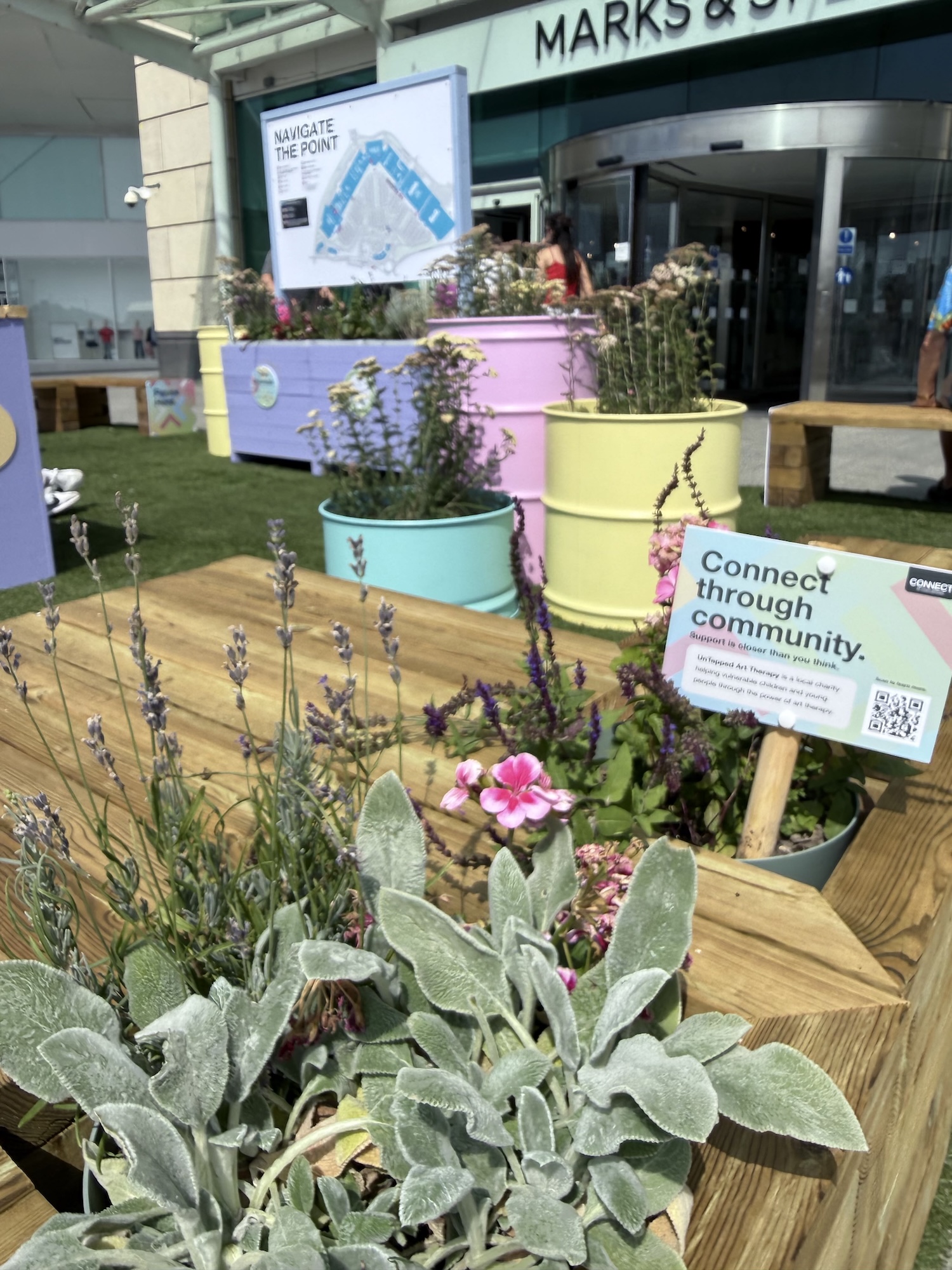 Potted flowers and silver-green foliage arranged on a wooden display outside a Marks & Spencer store, with pastel-coloured planters and a sign reading 'Connect through community'.