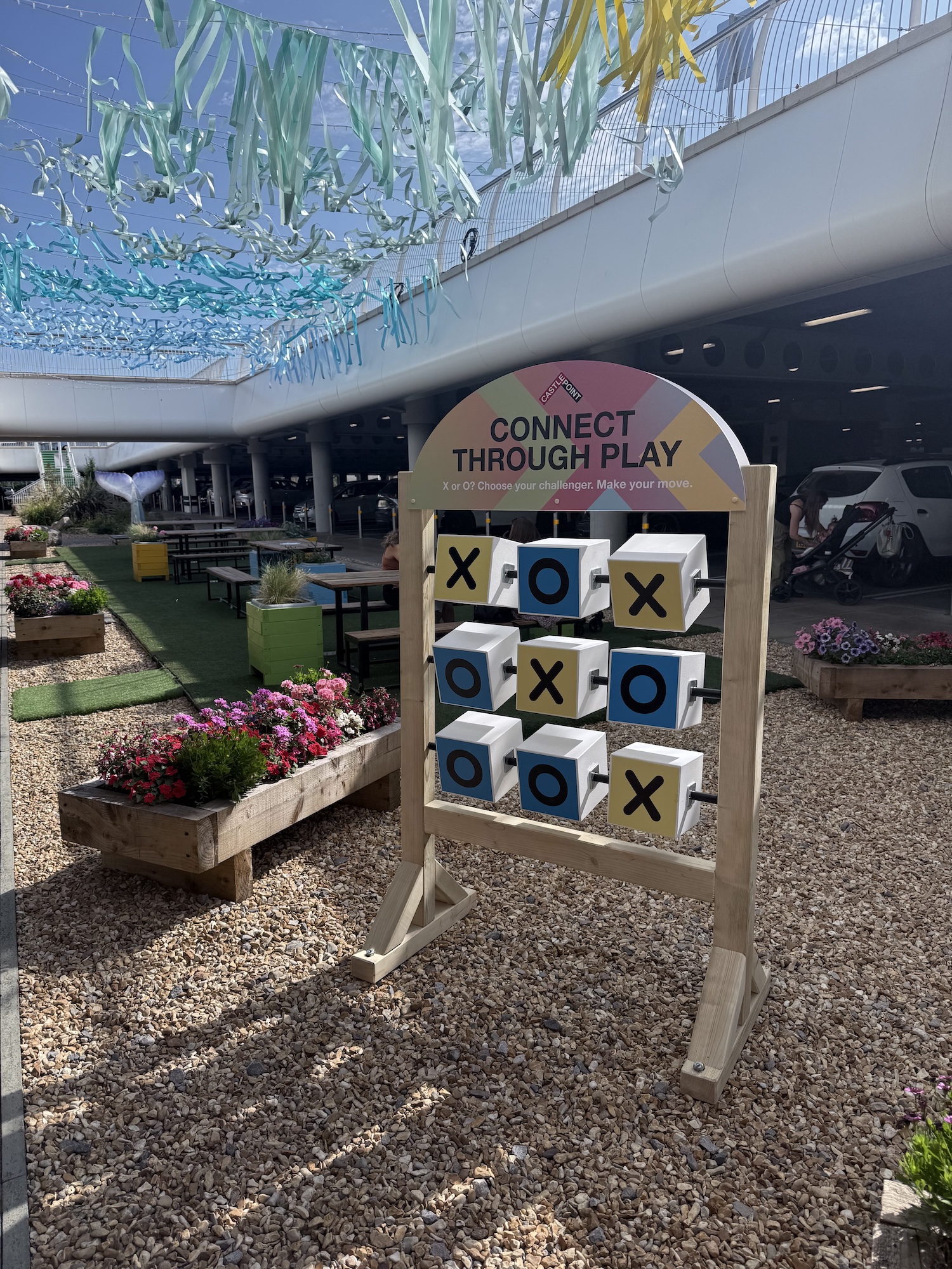 A freestanding Connect Through Play tic-tac-toe game with rotating cube blocks, set on a gravel patio beneath hanging streamers and beside wooden planters with flowers.