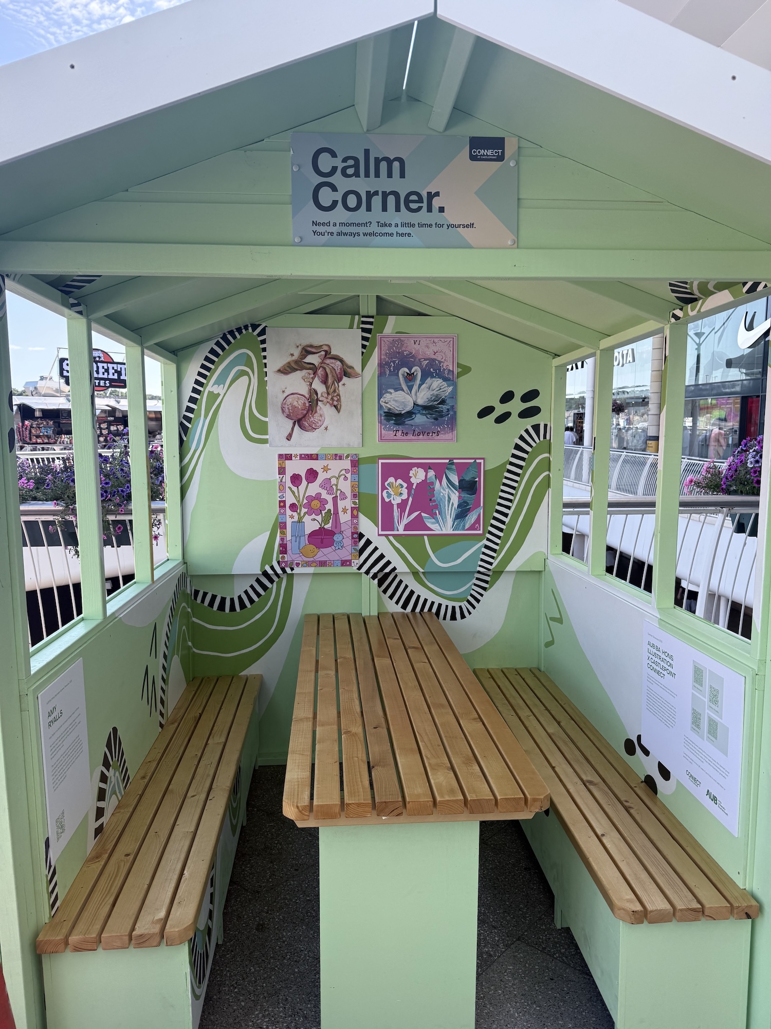 A mint-green outdoor shelter with wooden benches and a central table, decorated with colourful artwork and a sign reading Calm Corner.