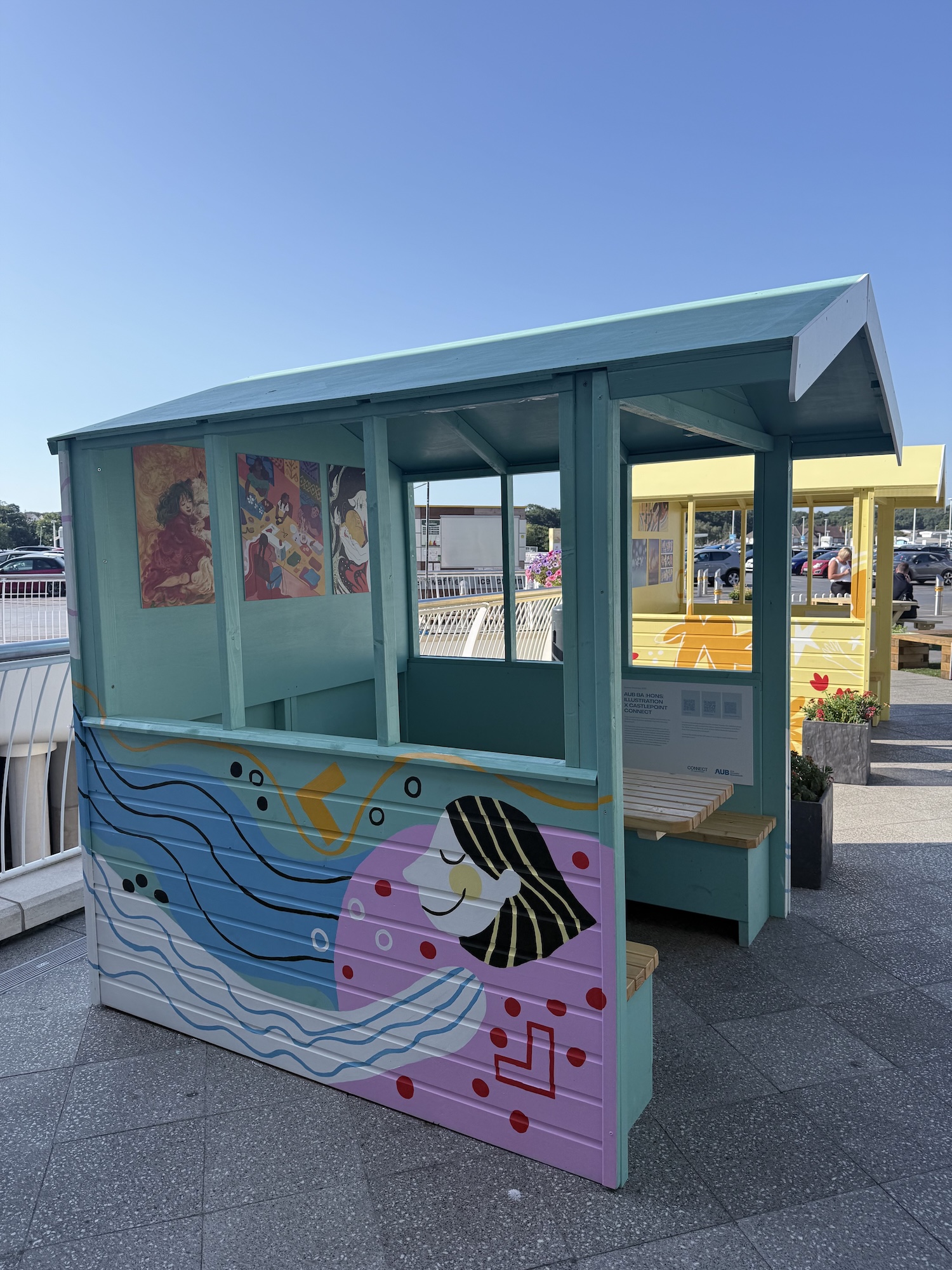 A turquoise painted kiosk with a patterned front panel and open side windows sits on a paved waterfront area, with another yellow kiosk behind it.