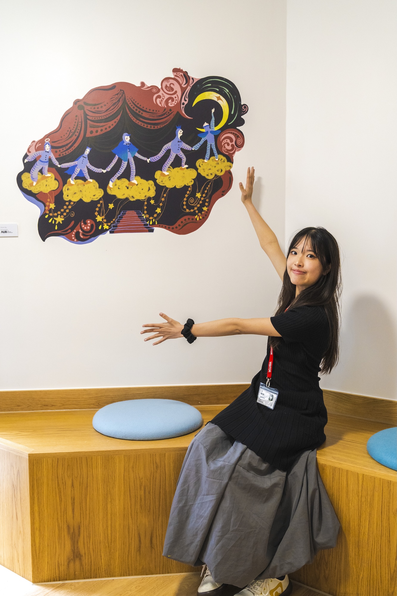 BA (Hons) Illustration student C Ng sitting next to and presenting towards a colourful vinyl illustration on the wall next to them.