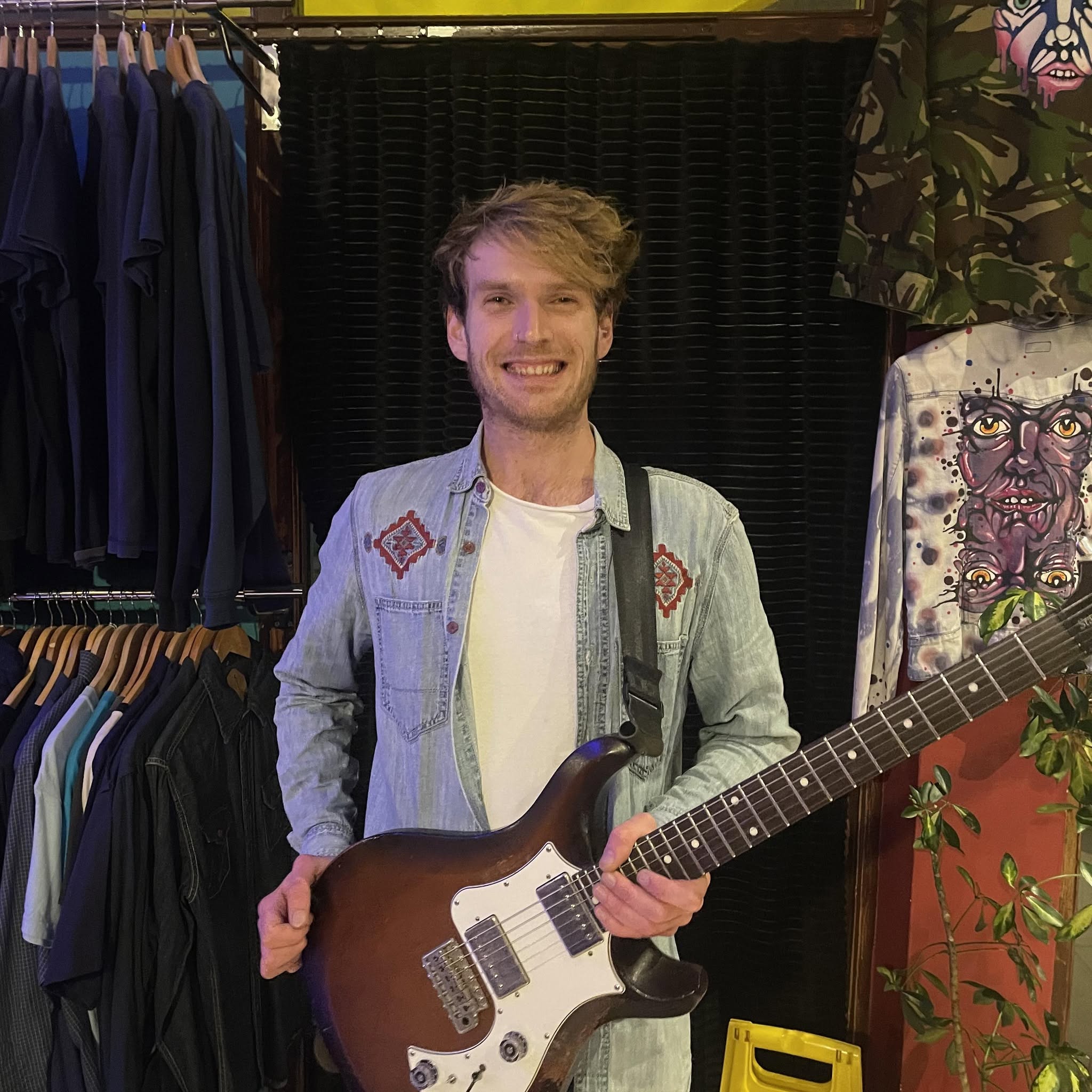 Musician C Ryan holding a guitar, standing between two rails of clothes.