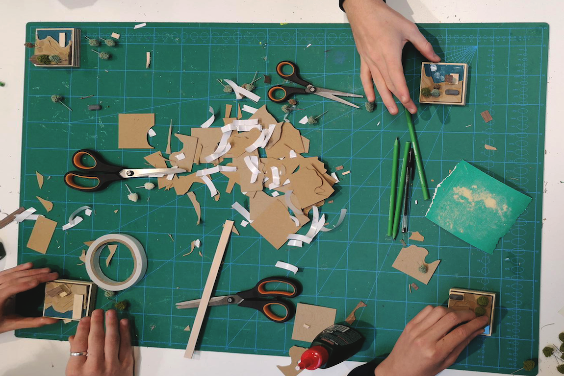A large cutting board in the middle of a table with lots of cut-up card and paper, pairs of scissors and tape.