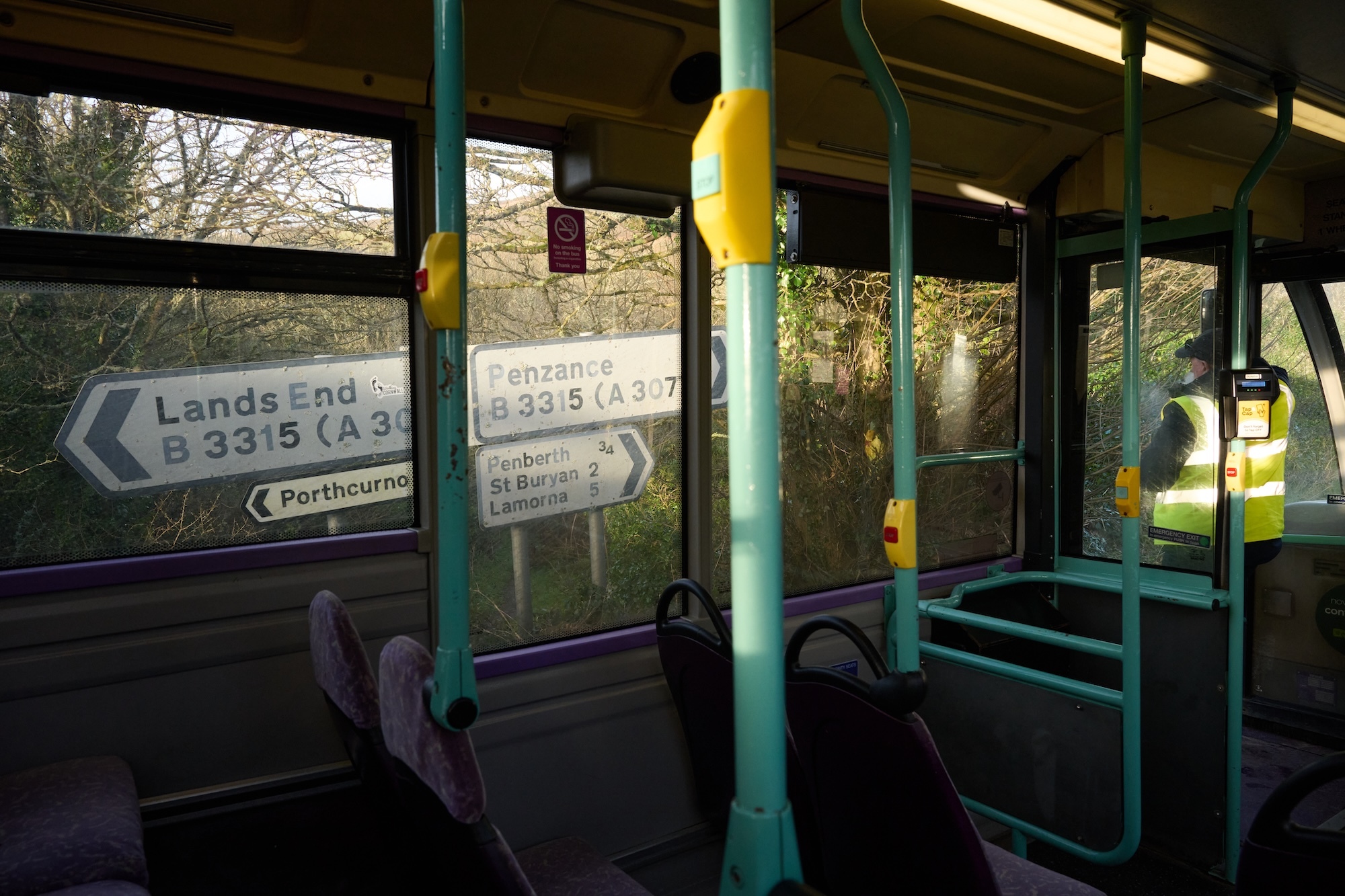Bus interior with teal handrails and seat frames, purple upholstery. Windows show countryside road signs including "Lands End B3315".