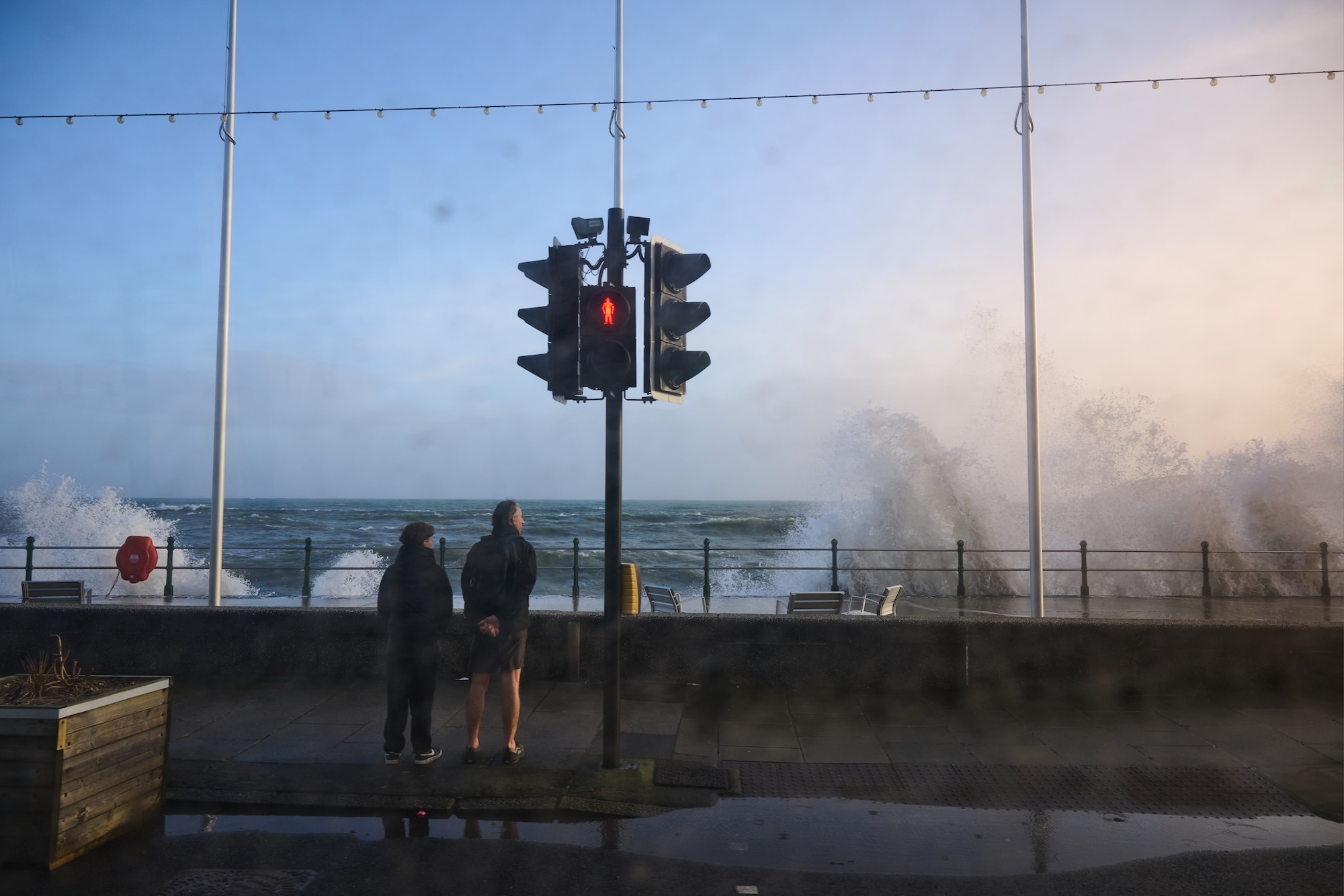 Traffic lights on red with two people standing nearby as large waves crash over seafront railings, creating dramatic spray against blue sky.